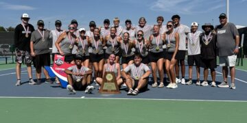Group of young tennis players and coaches wearing medals, posing with a trophy on a sunny tennis court.