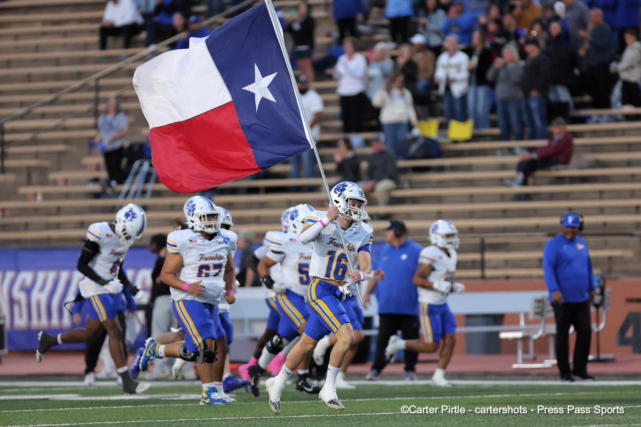 Football players in white uniforms run onto the field as a large Texas flag waves overhead, stands full of spectators in the background.