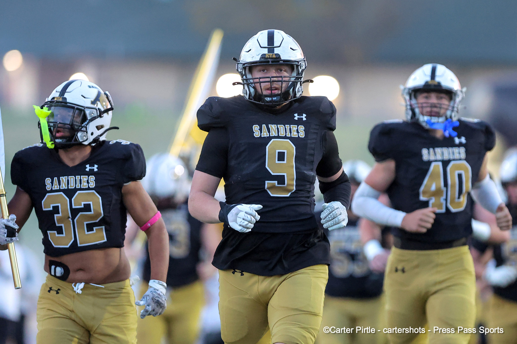 Three football players in black and gold Sandies uniforms sprint onto the field, center player wearing number 9 with a white helmet.