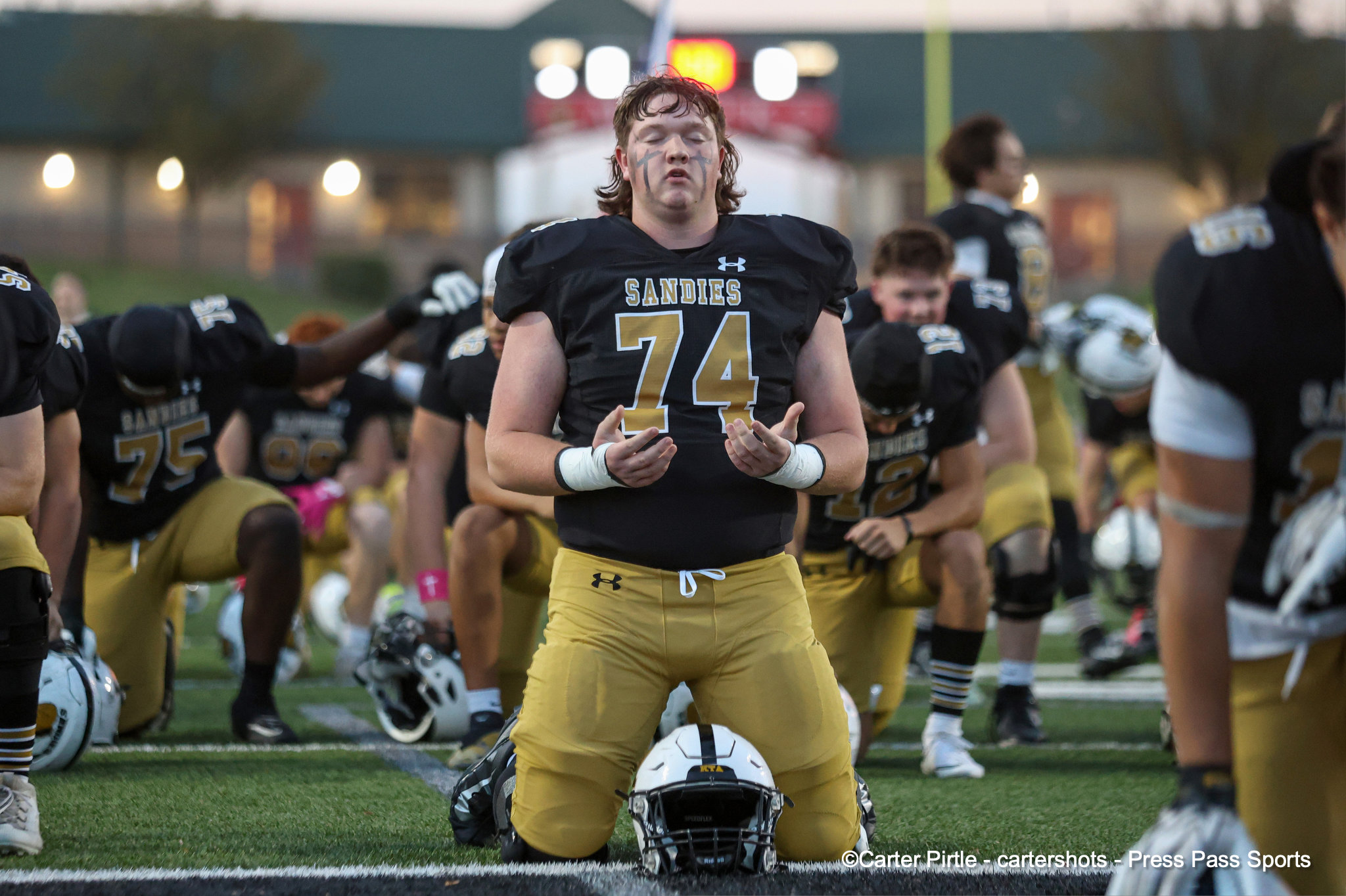 Football players kneeling on the field, central player wearing black jersey with'Sandies' and number 74 amid teammates.