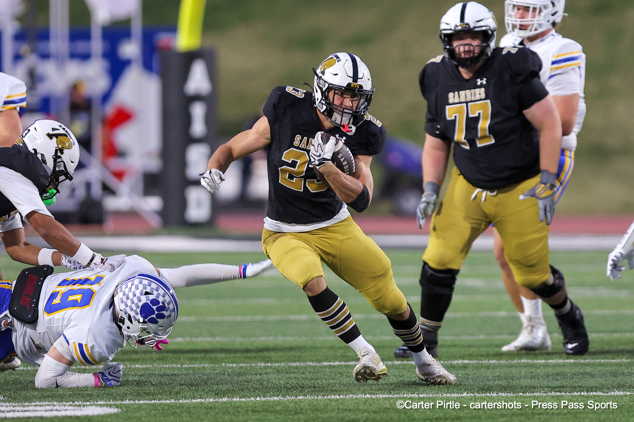 Sandies running back (black jersey, gold numbers) carries the ball as a tackler on the ground and a pursuing defender in white and gold closes in on a green football field.