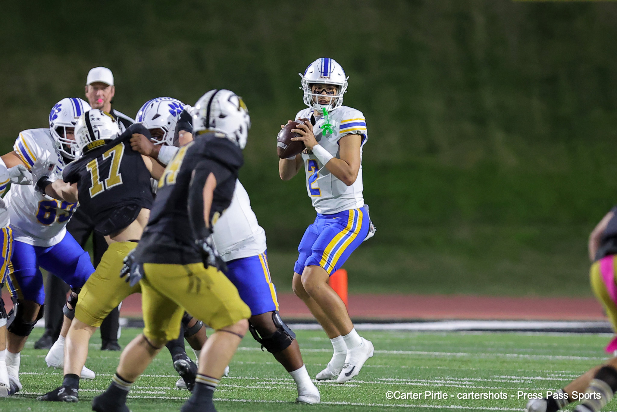 Quarterback in a white and blue uniform crouches to throw, surrounded by opposing players on a football field during a play.