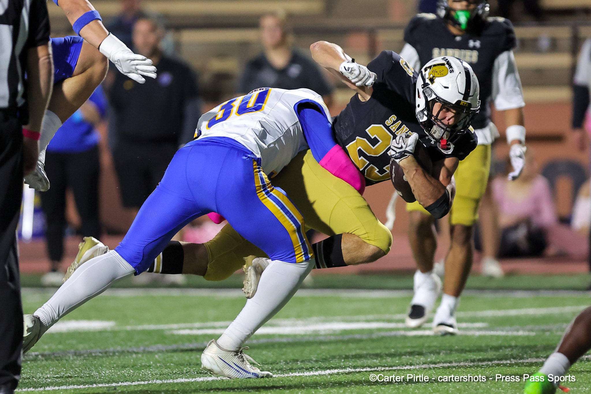 Two football players collide in a tackle on a green artificial turf field during a game, one in white-blue and the other in black-gold.