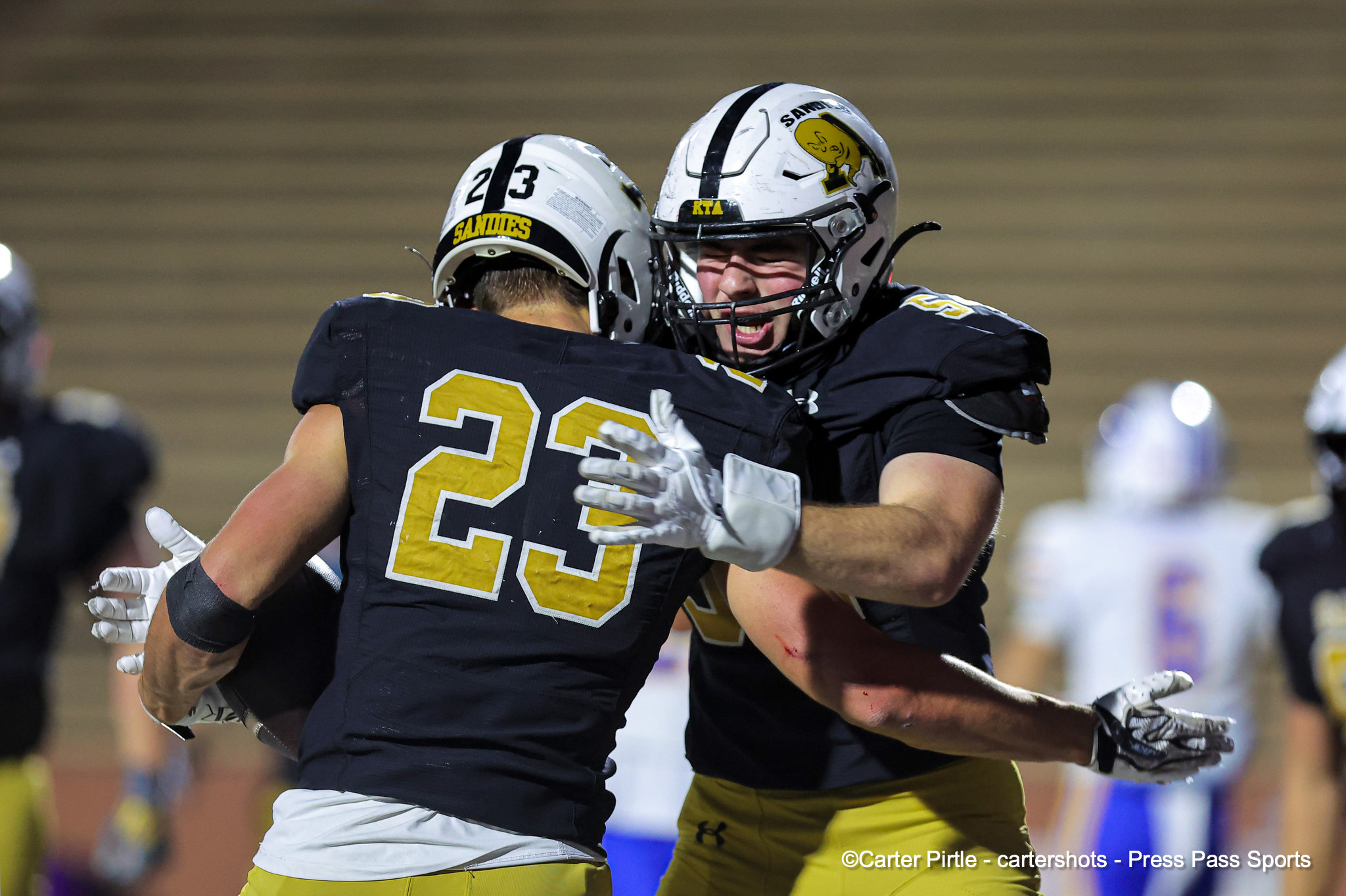 Two football players in black jerseys hug each other in celebration on the field, numbers visible (23).