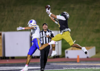 20261022-Frenship-AHS-FB-011 | Press Pass Sports Two football players jump for a pass in midair; one in white/blue and the other in black/yellow, with a referee nearby on a night game field.
