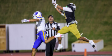 Two football players jump for a pass in midair; one in white/blue and the other in black/yellow, with a referee nearby on a night game field.