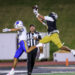 20261022-Frenship-AHS-FB-011 | Press Pass Sports Two football players jump for a pass in midair; one in white/blue and the other in black/yellow, with a referee nearby on a night game field.