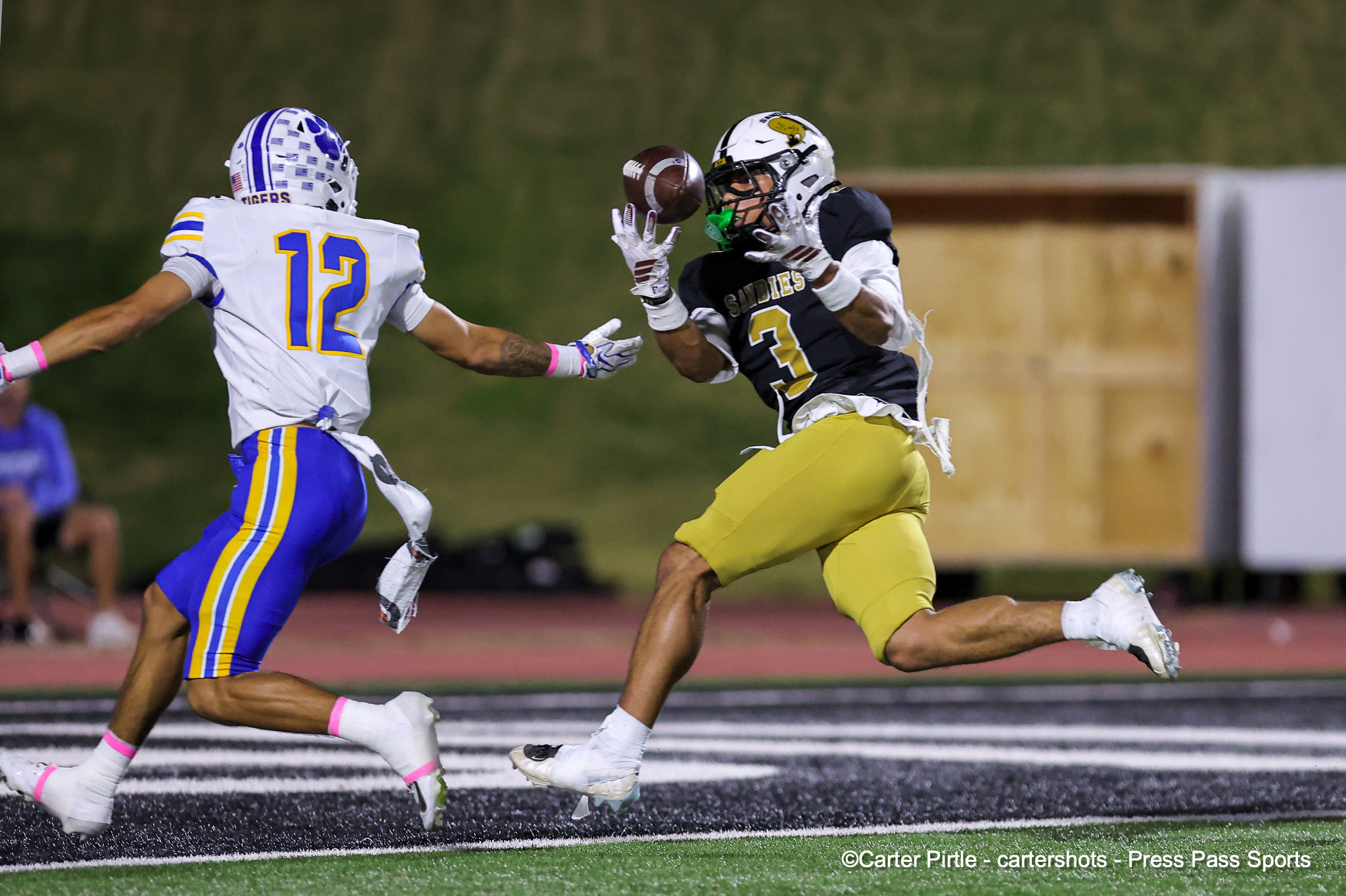 Two football players leap for the ball mid‑play; white‑and‑purple team on left, black and gold on right, on a turf field.