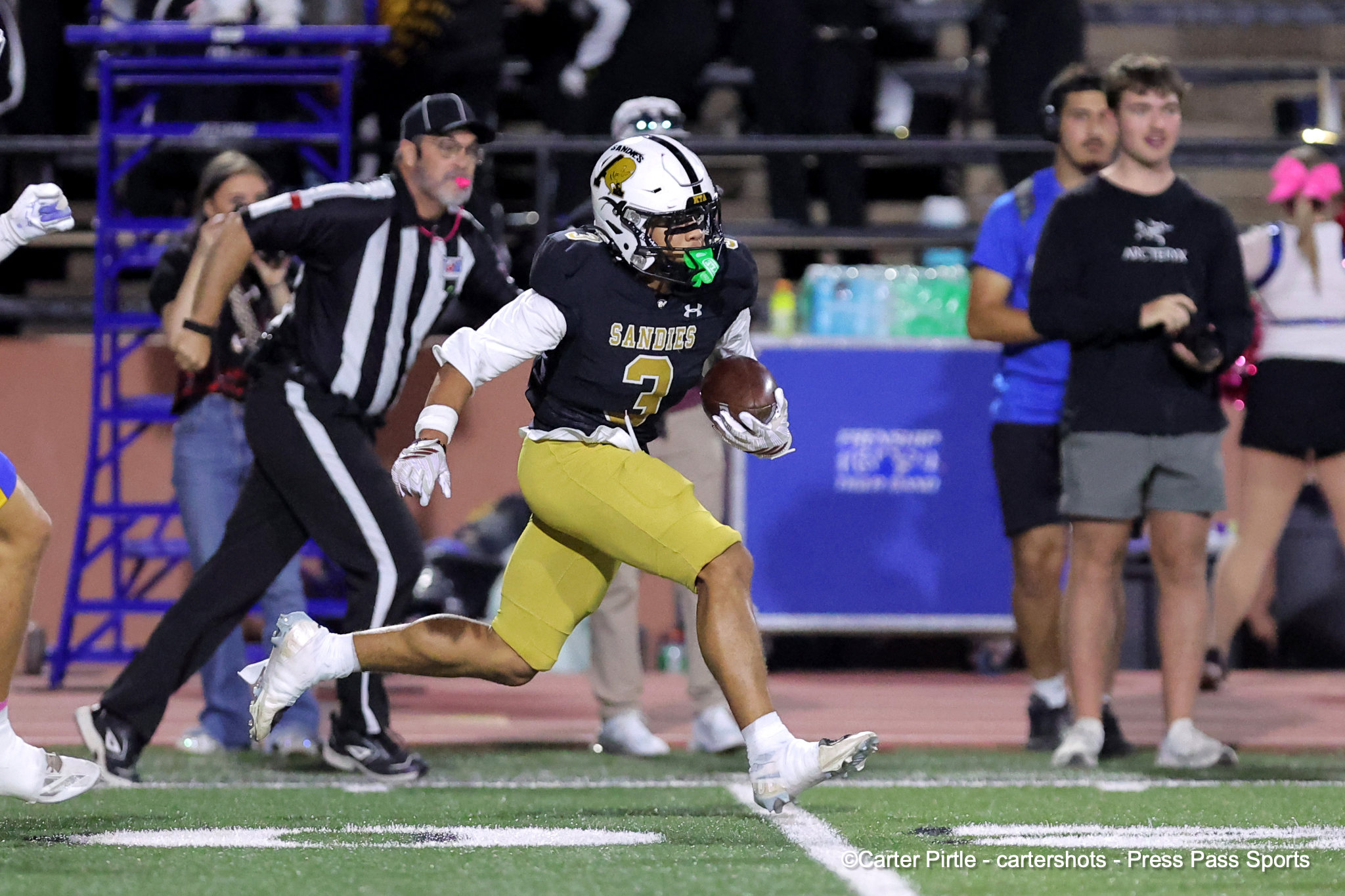 Sandies player wearing number 3, white helmet and black jersey, sprinting with the football down the sideline as officials and defenders pursue nearby on a football field.