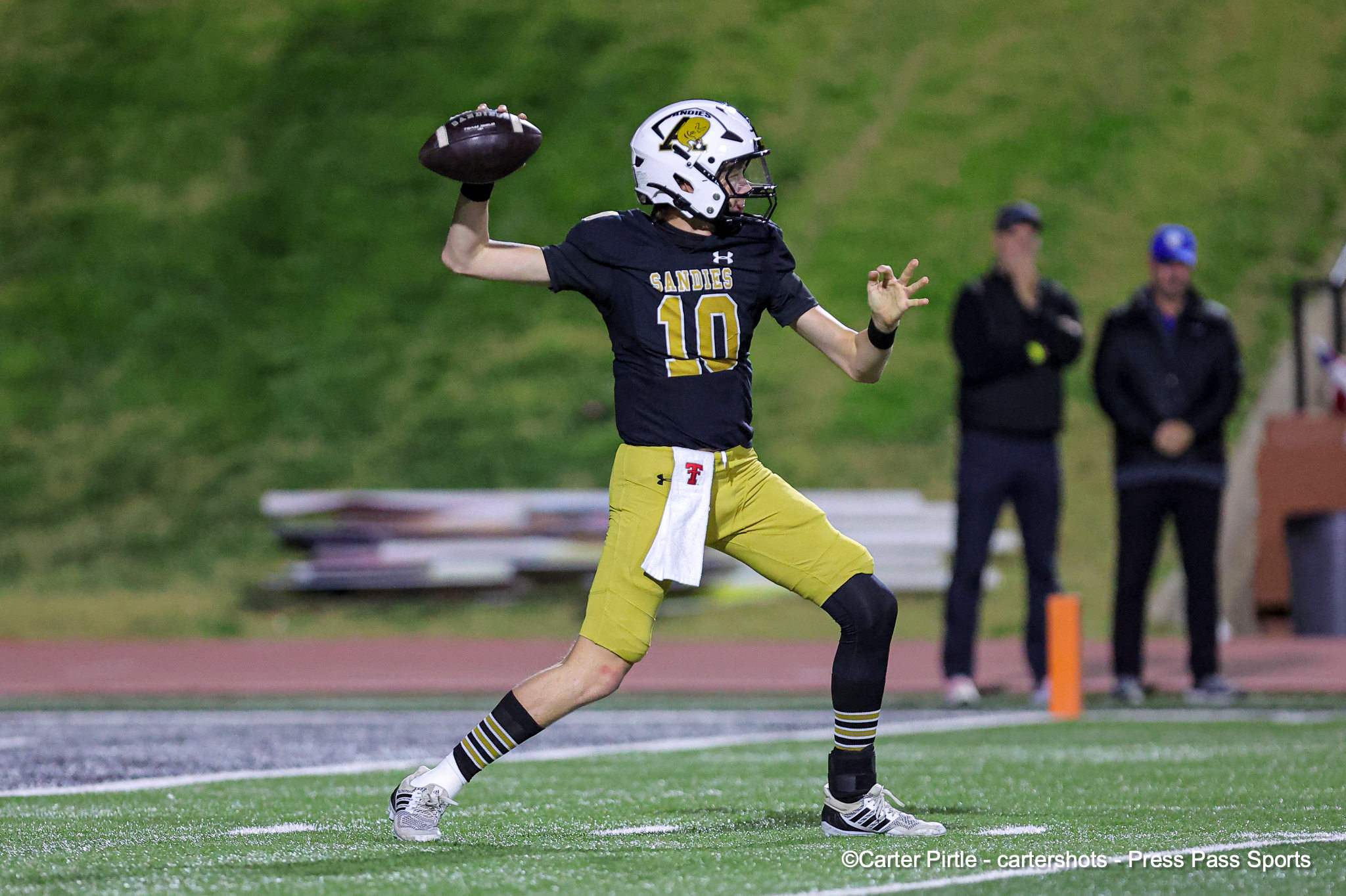 Sandies football player #10 in black and gold, white helmet, running with the ball and preparing to throw on a green field. In the background, spectators stand by the sideline.