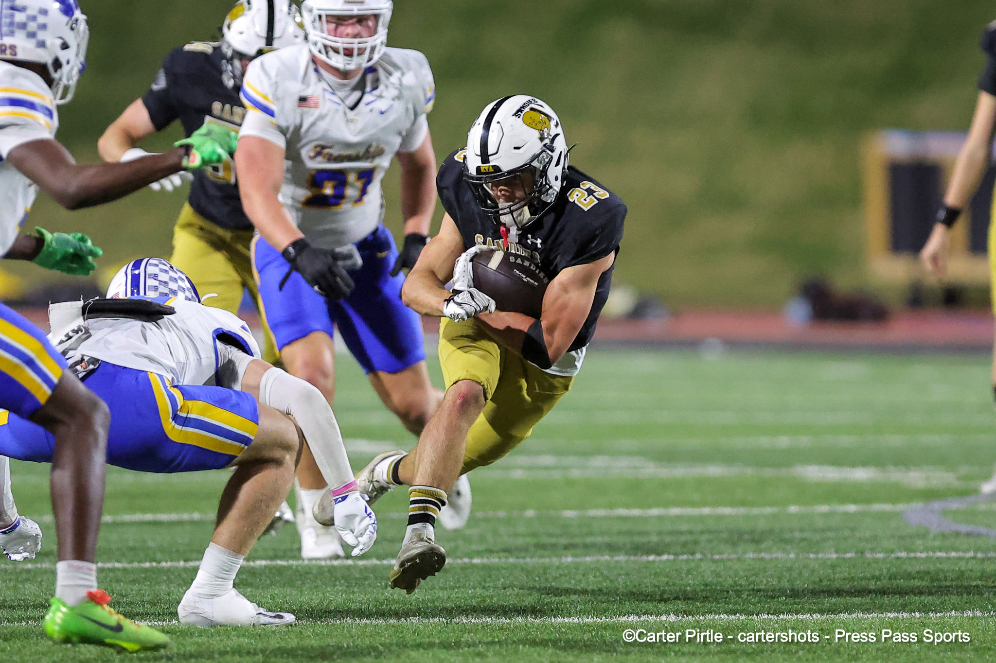 Football player #23 in black jersey carries the ball as defenders in white/blue close in on a green field fieldside tacklers nearby.