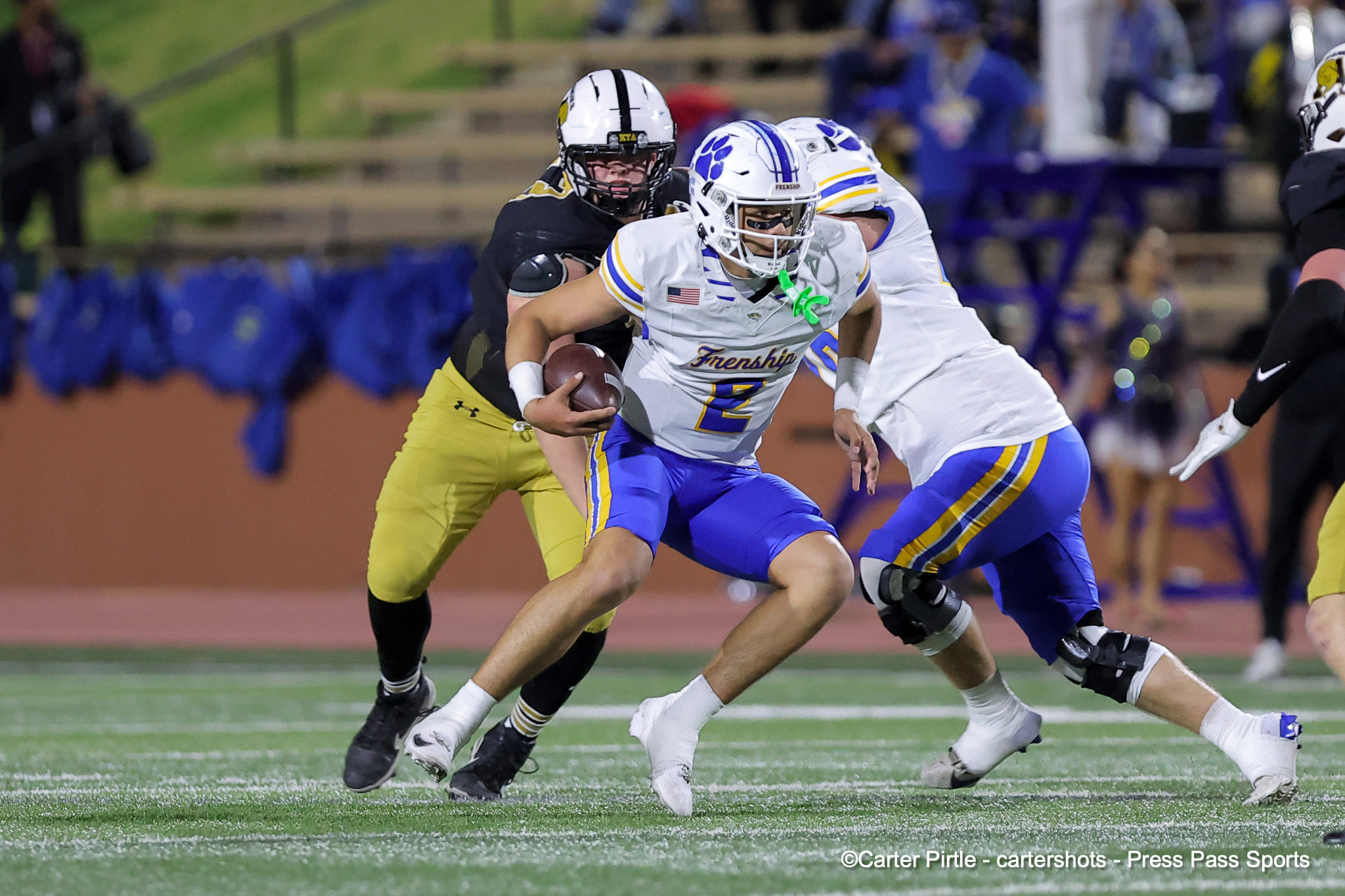 Football action: a white-uniformed runner with the ball is pursued by a defender in black and gold on a green turf field with blurred stands in the background.