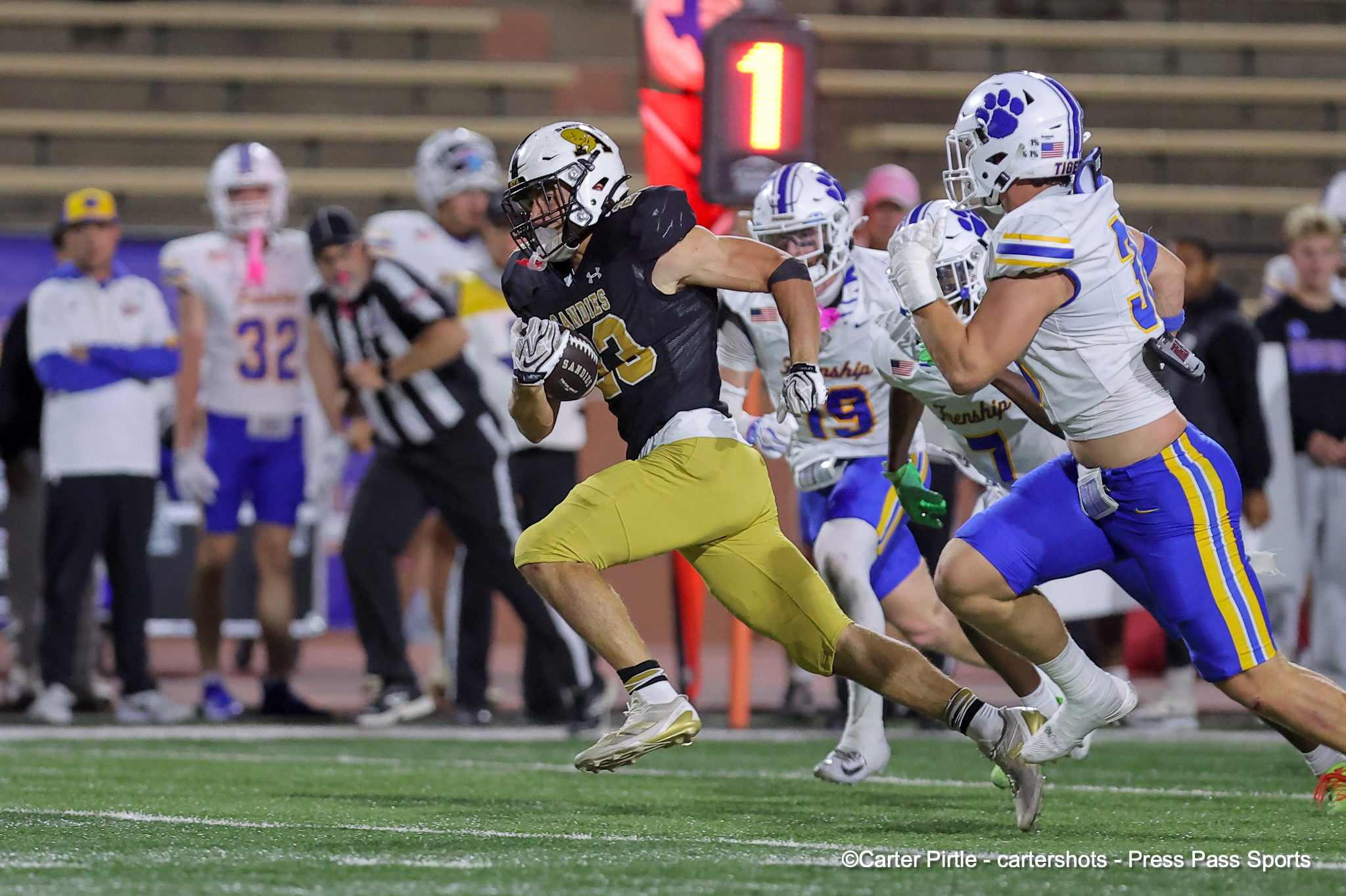 Football players sprinting down the field; a ball carrier in black/gold dodges defenders in white/purple uniforms.