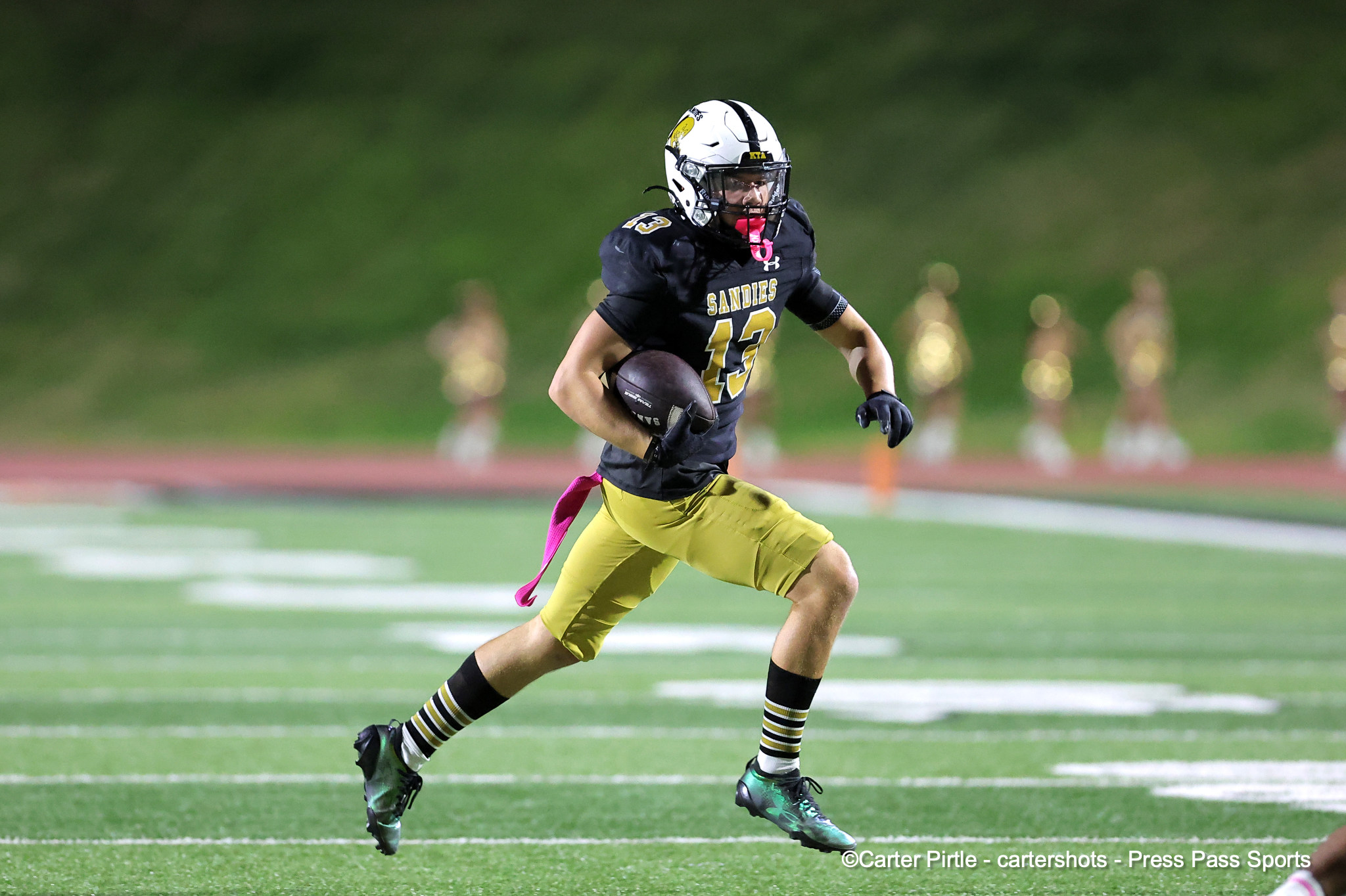 Young football player in black-and-gold uniform carrying a ball on a green turf field, pink flag visible at waist.