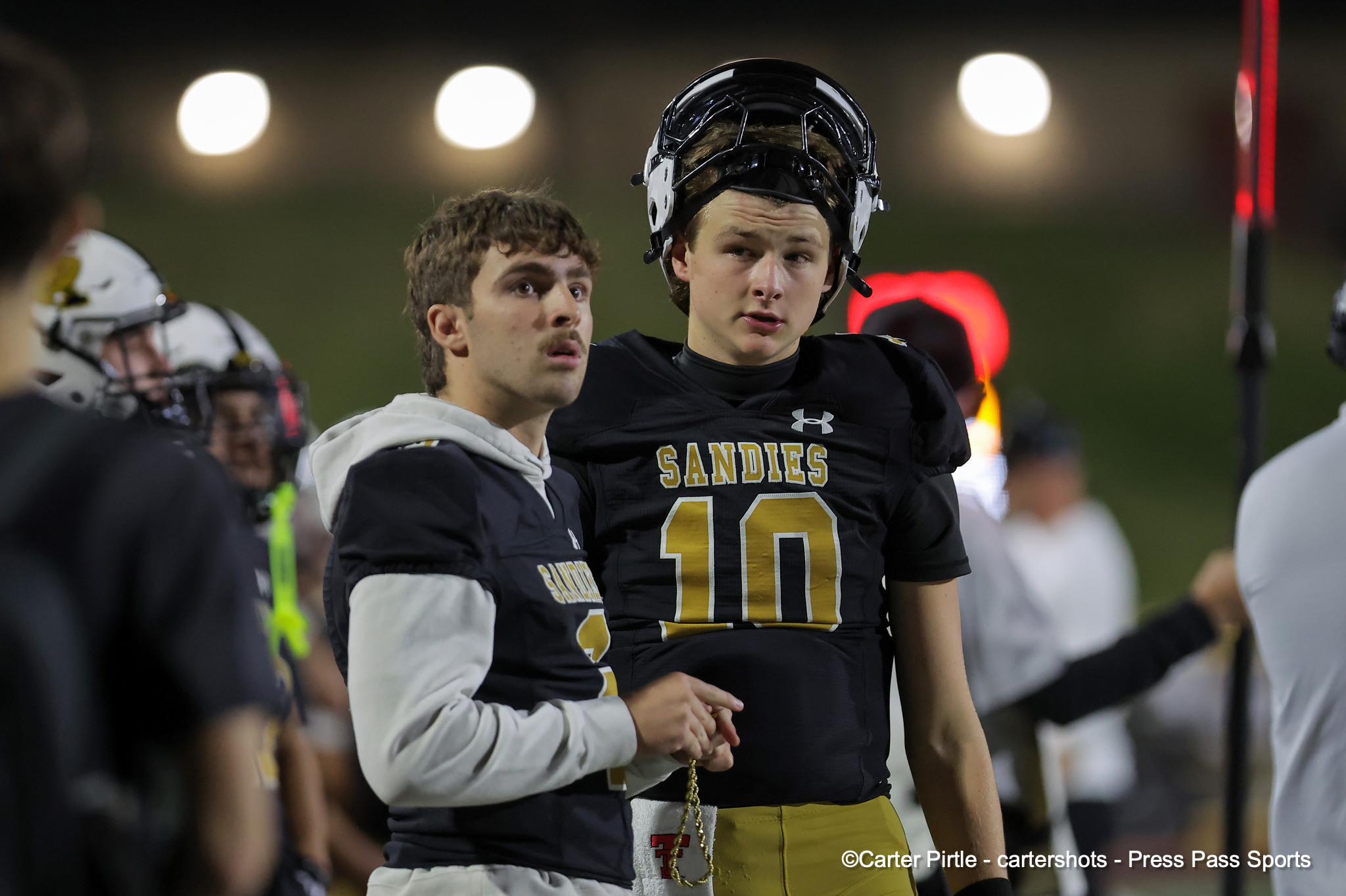 Two high school football players in black and gold Sandies uniforms on the sideline, one with helmet on, #16 visible on the jersey.