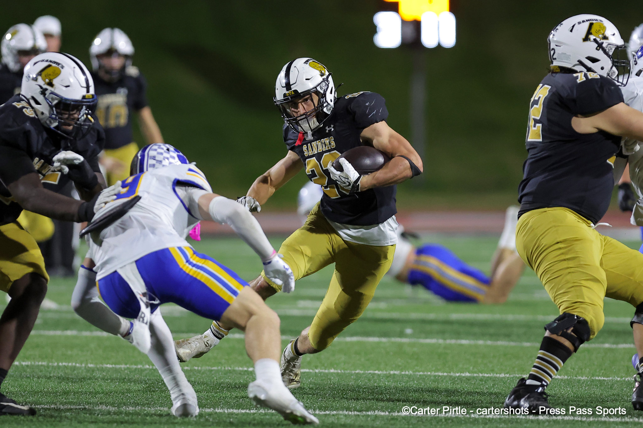 Football player in black and gold carries the ball toward the sideline while defenders close in during a night game on a turf field.