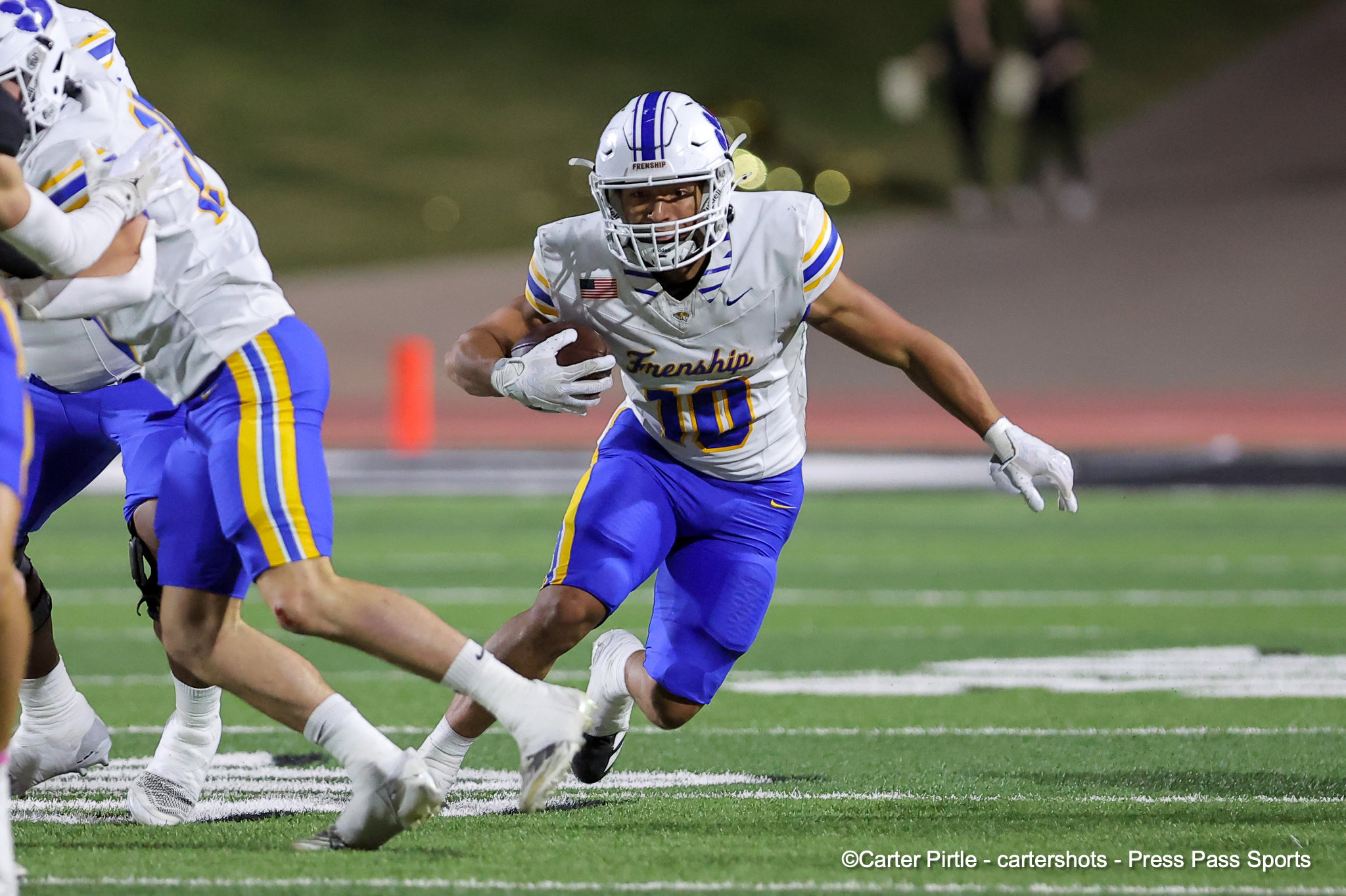 Football player in white and blue with the ball, sprinting downfield while a defender closes in on a green field at night.