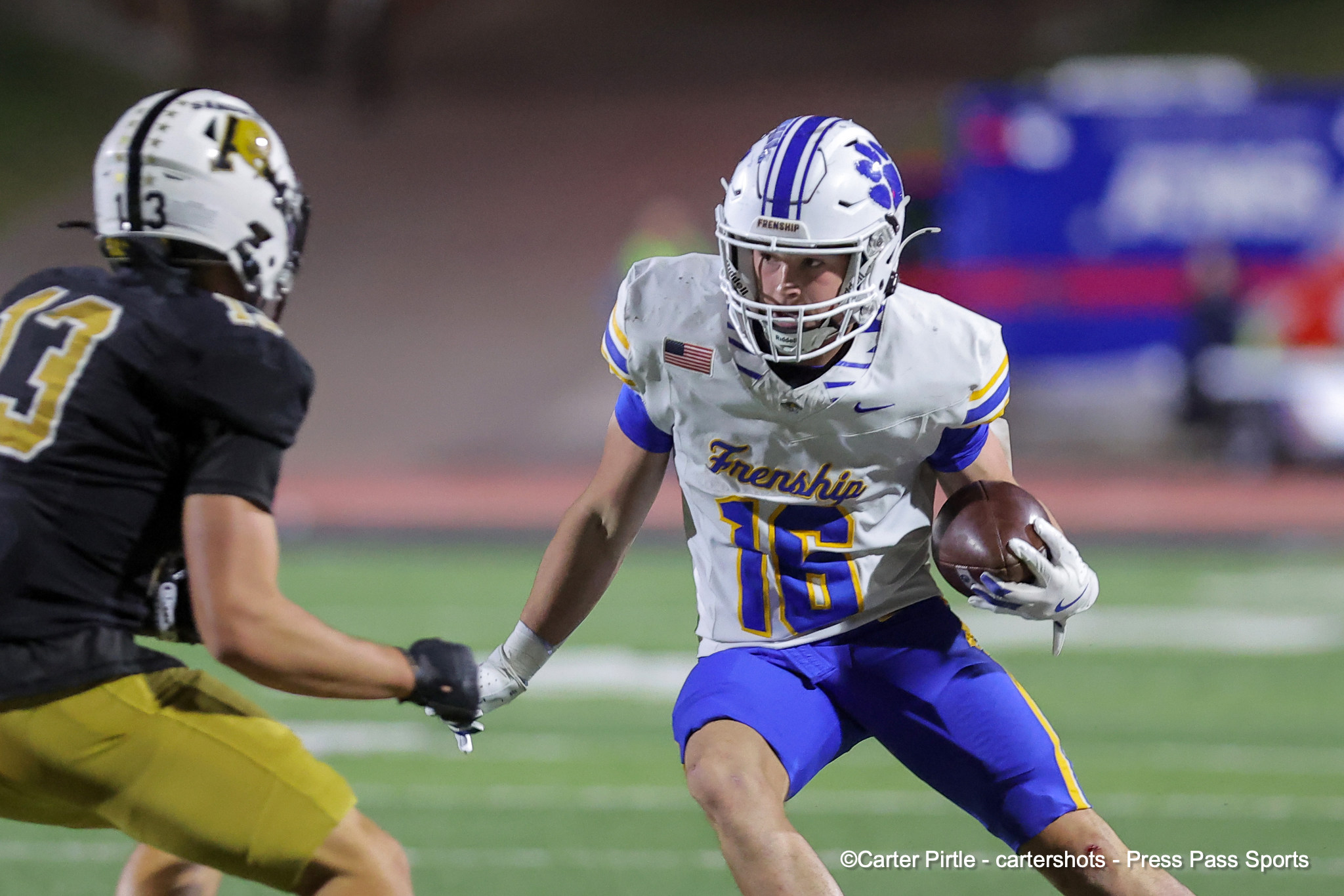 Football player in white and blue with ball powers forward as a tackler in black and yellow approaches on a grassy field.