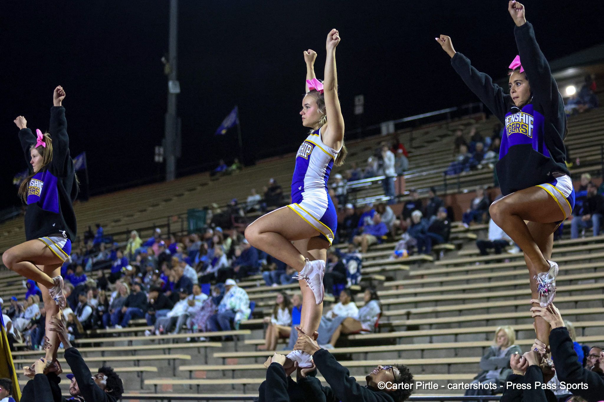 Cheerleaders perform a high-rise stunt on stadium bleachers at night, flyers held up by bases with pink bows in their hair.