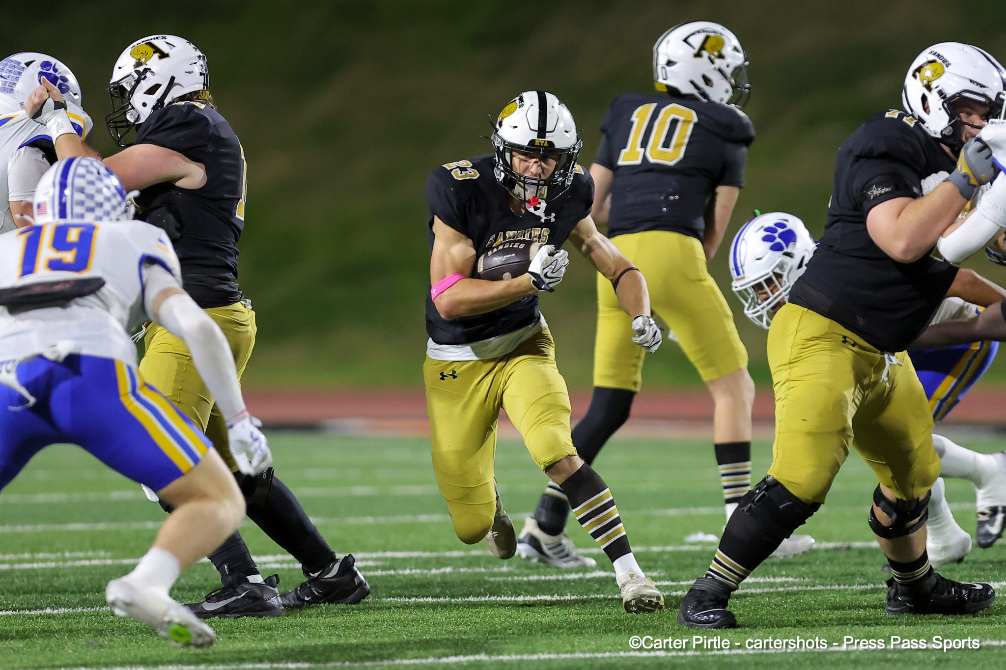 Young football player in black and gold jersey runs with the ball as teammates and defenders close in on a green turf field hop-scene style action shot.