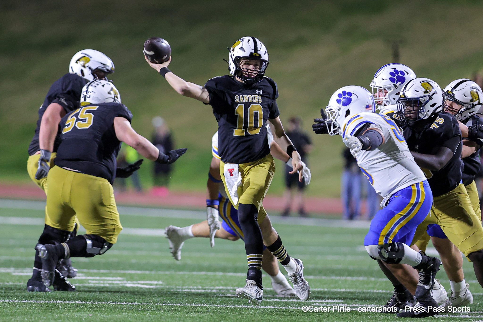 Quarterback in black and gold (#10) throws a pass as teammates and defenders close in on a crowded field scene in a football game.