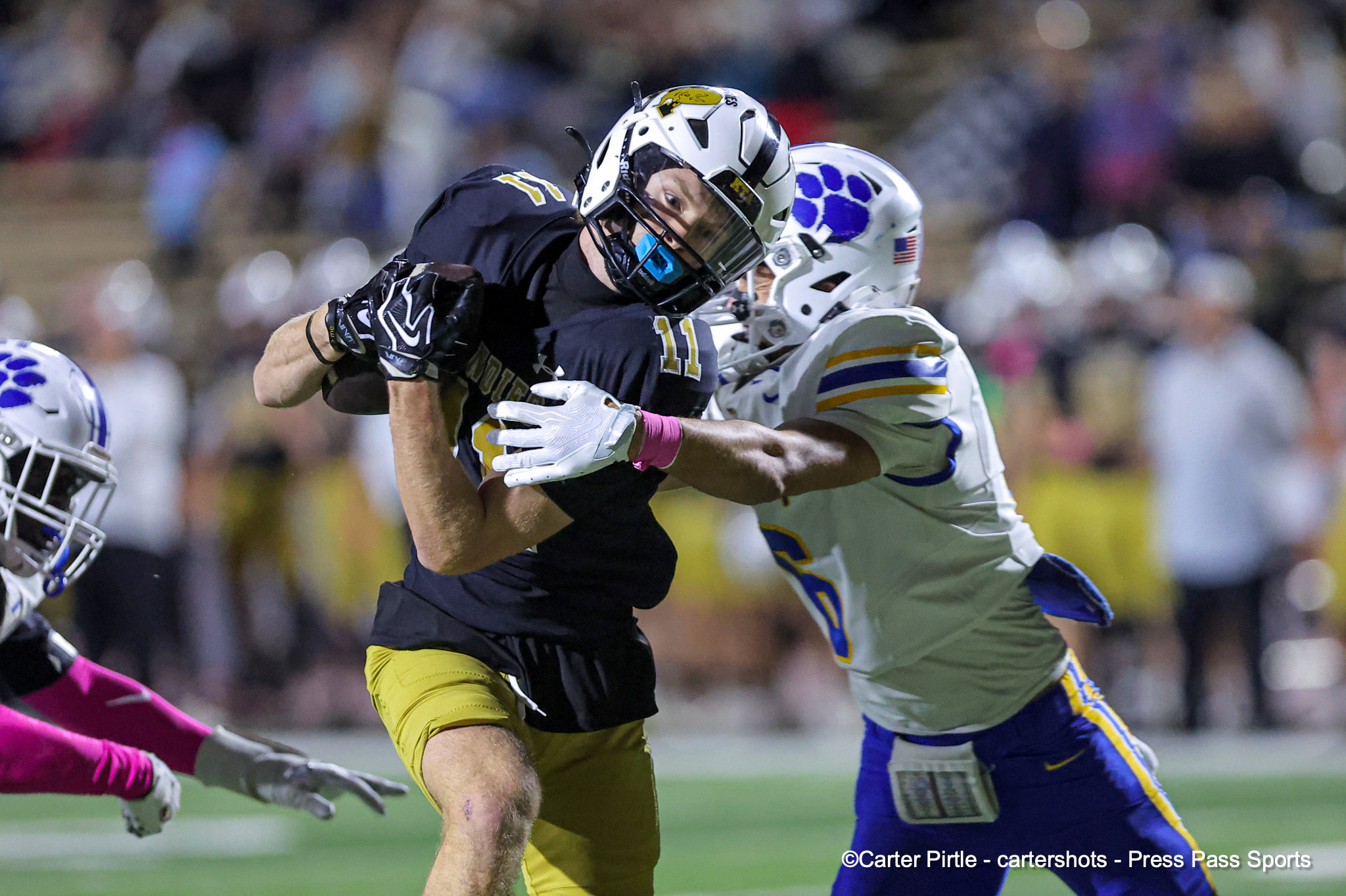 Two football players collide during a game, one in black and gold reaching for the ball while a white-uniform opponent with blue accents tackles from the side.