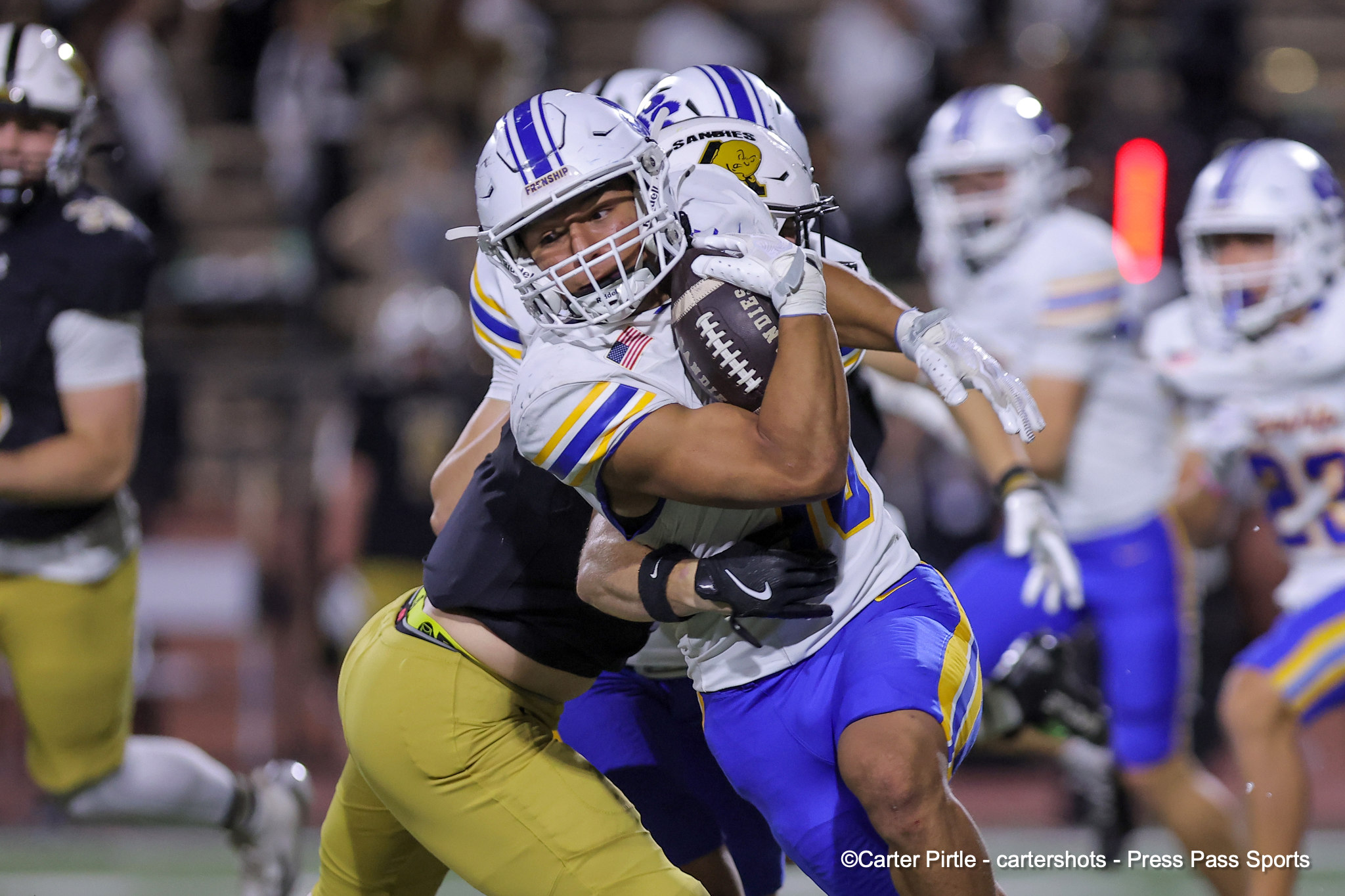 Two football players in white and blue jerseys grapple for the ball during a game, crowd blurred in the background.