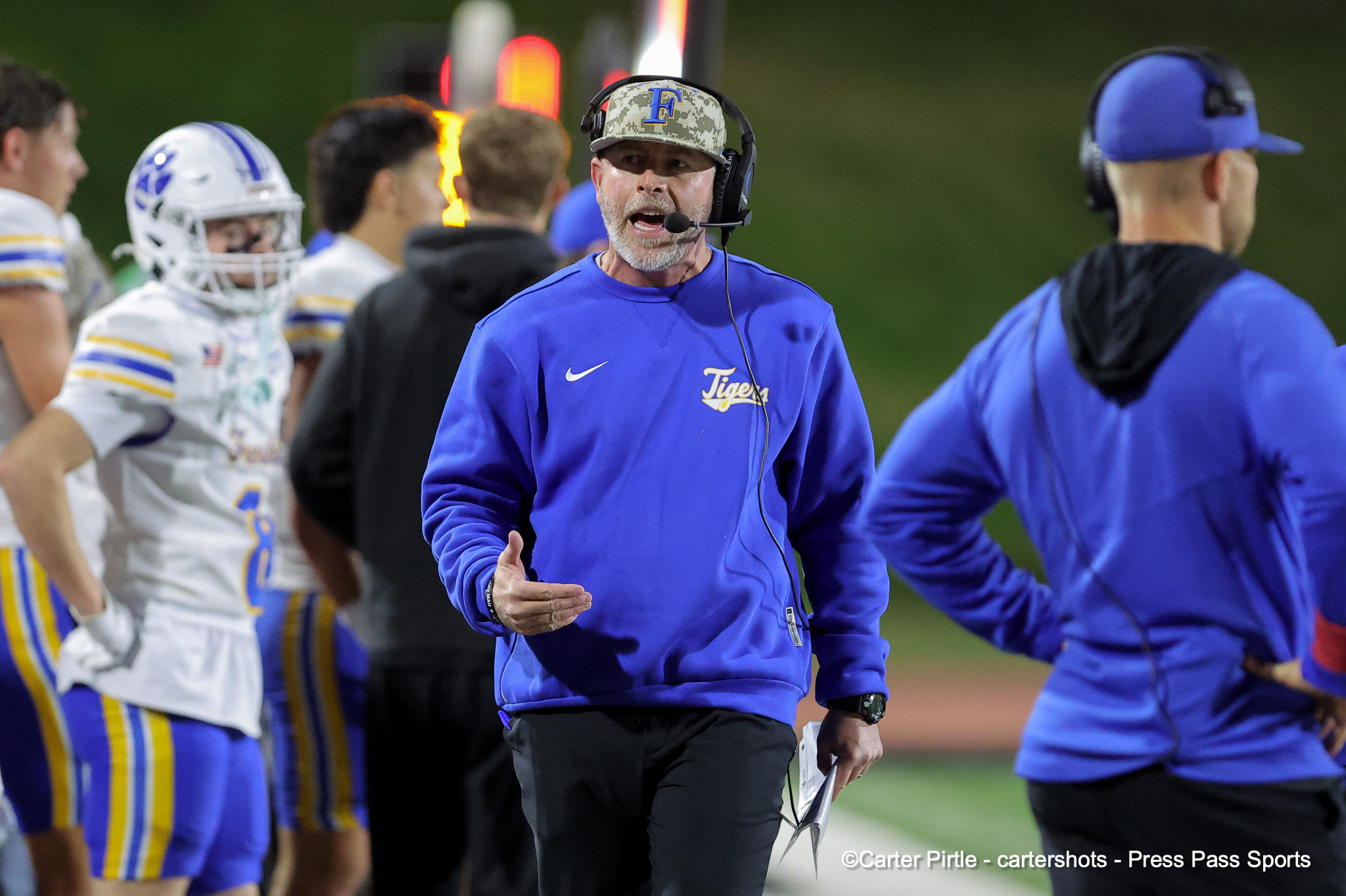 Football coach in a blue pullover with headset speaks to players on the sideline during a football game.