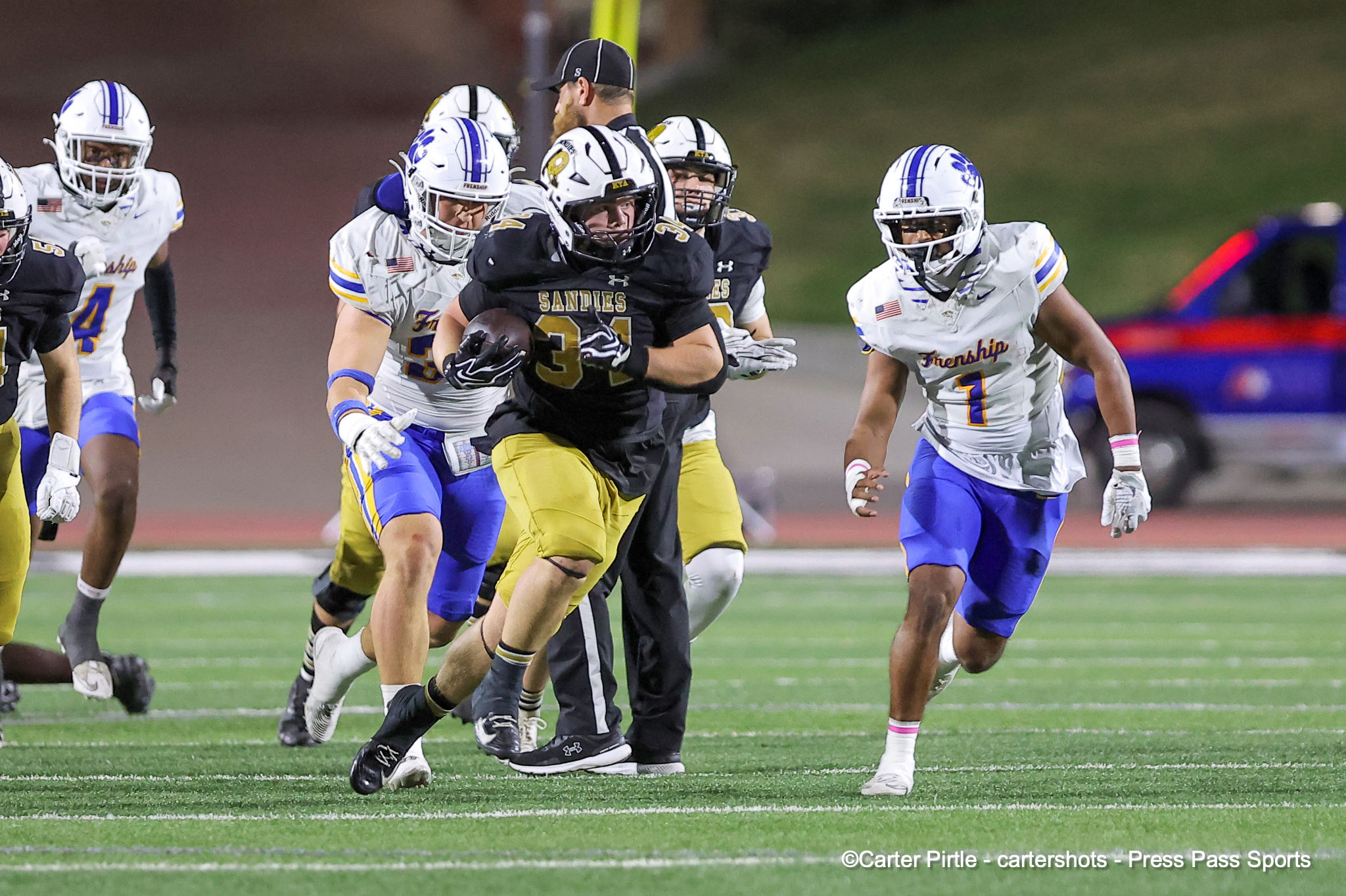Sandies player in black and gold carries the ball while defenders in white chase him on a green football field.