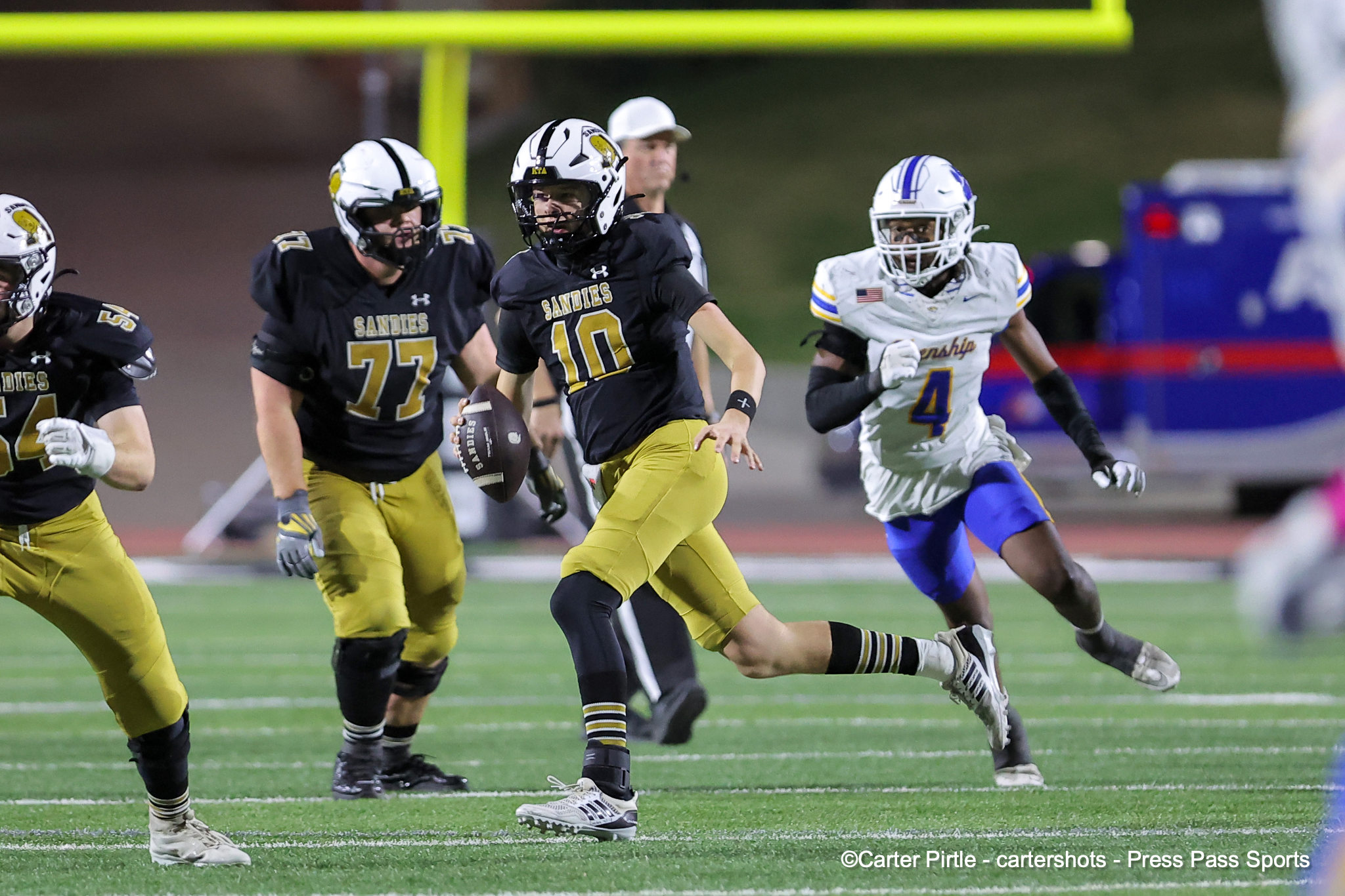 Sandies running back in black and gold (#10) carries the ball, pursued by a defender in white during a nighttime game distance shot.