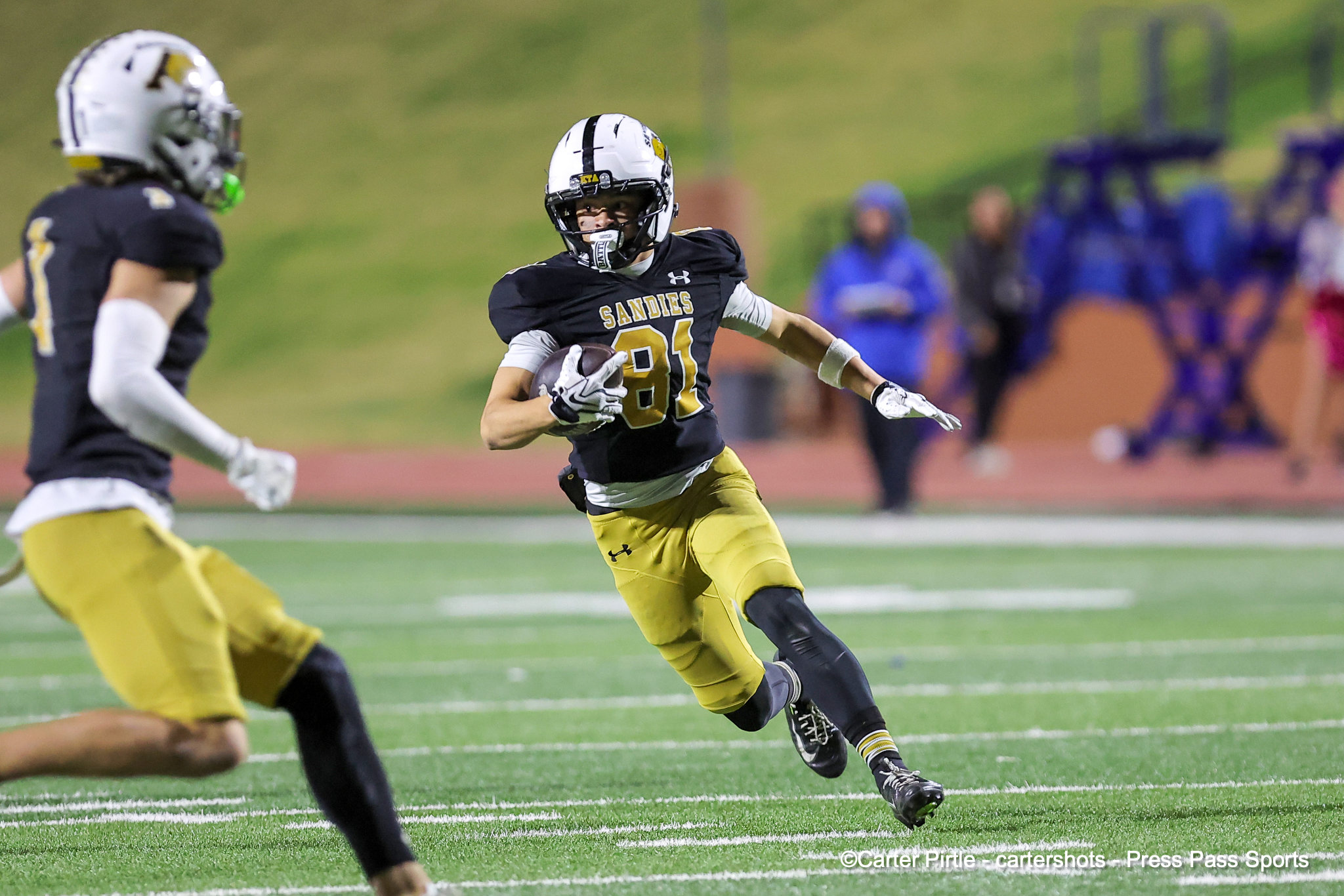 Football player wearing black and gold jersey with the ball, sprinting on a green field during a game.