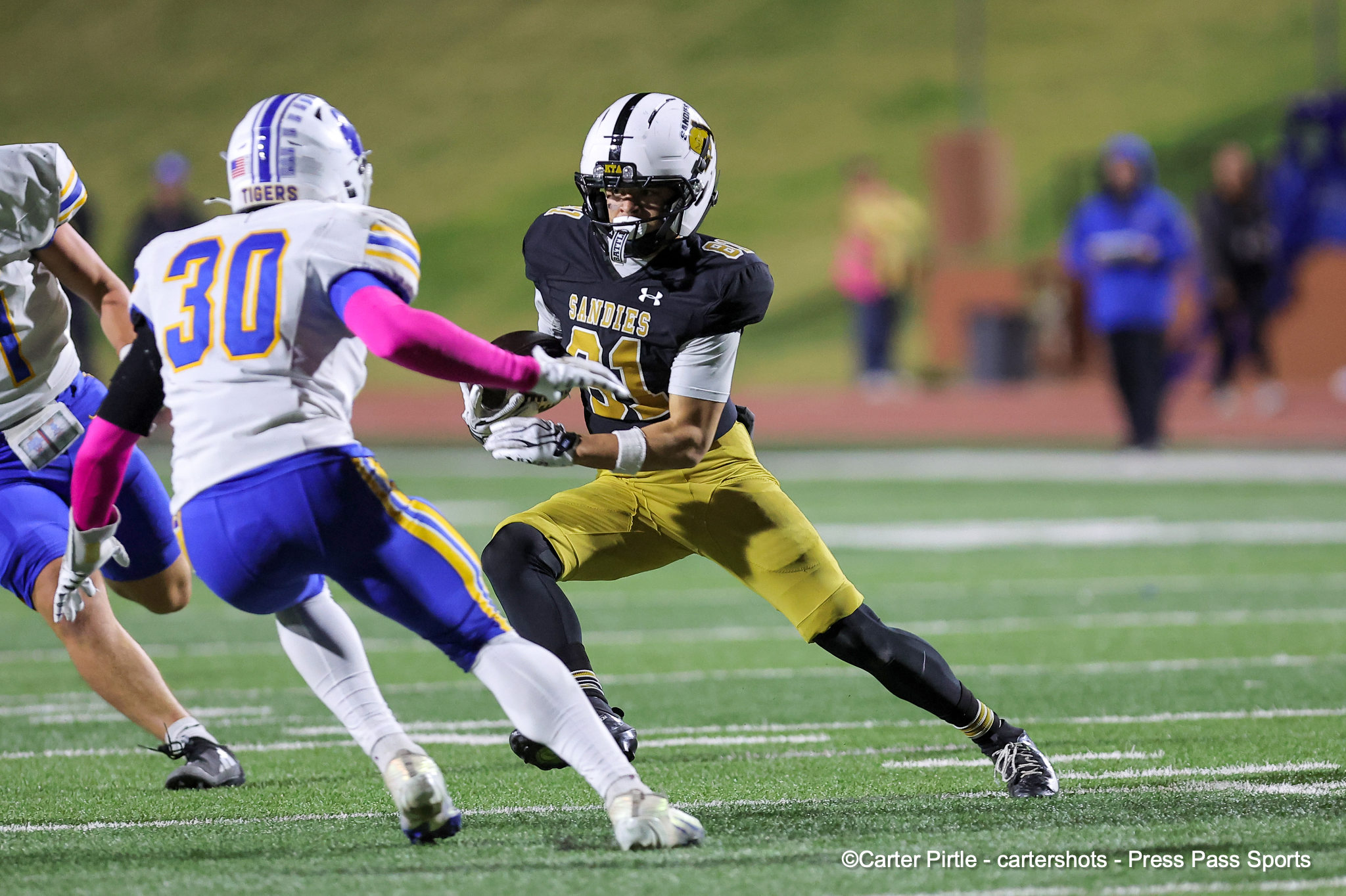 American football action: a Sandies defender in black jersey runs with the ball as teammates and opponents pursue on a green turf field.