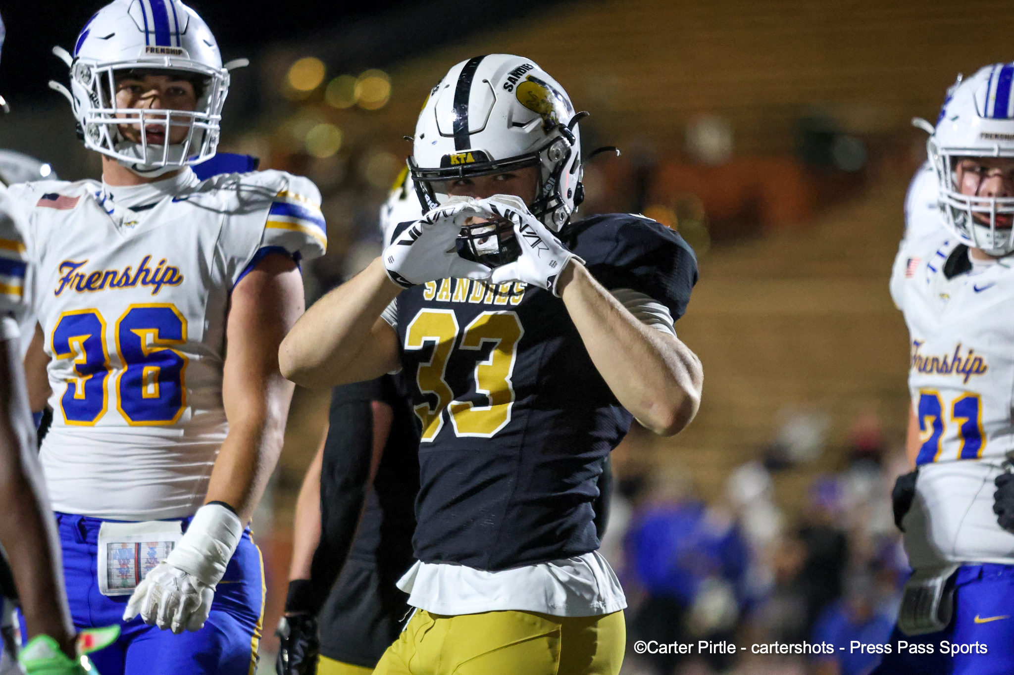 Nighttime football scene with players in white and black uniforms; central player in black makes a hand gesture in front of his chest while teammates look on.