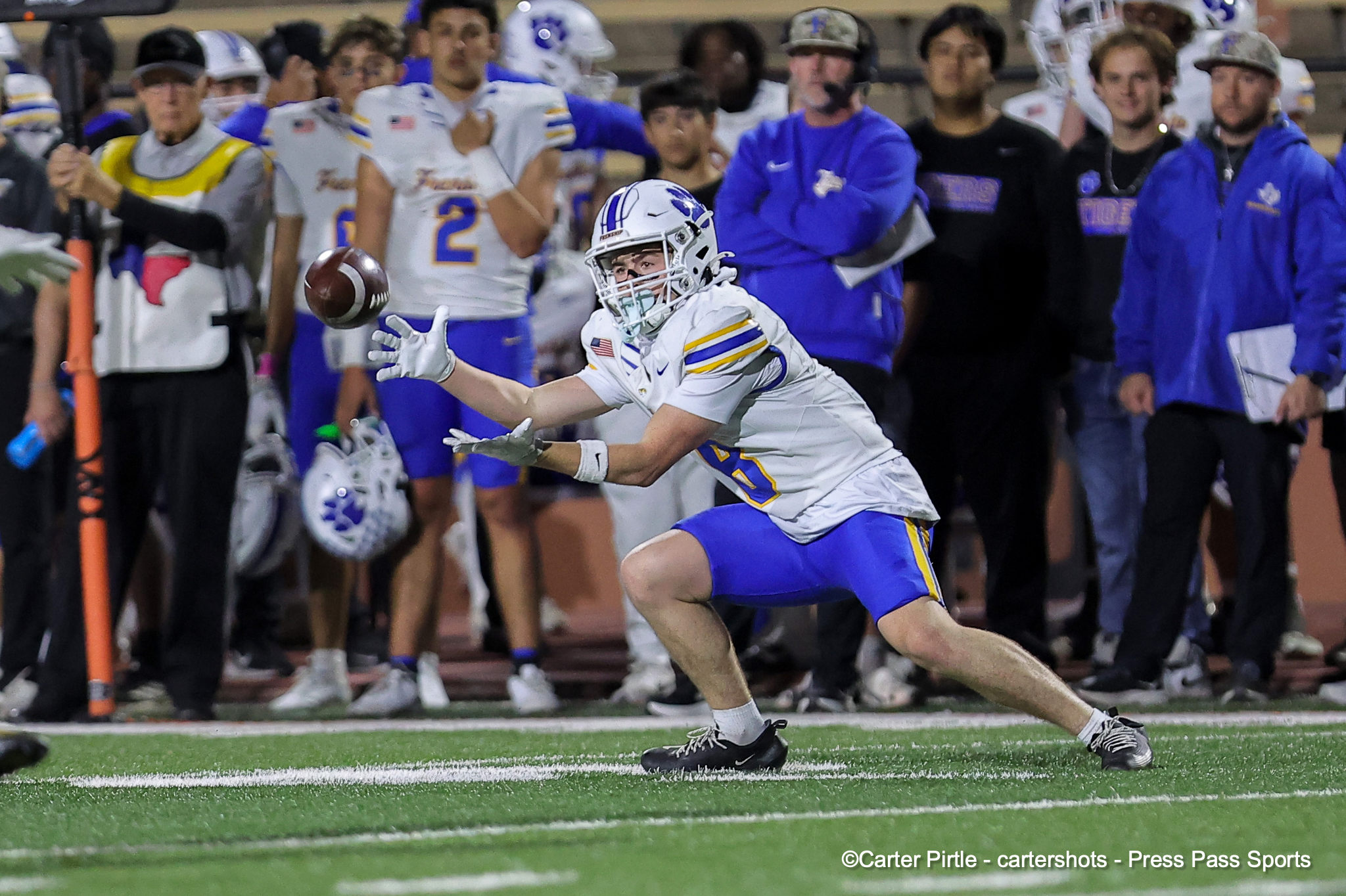 Football player in white and blue lunges for a loose ball near the sideline as fans watch from the sidelines.