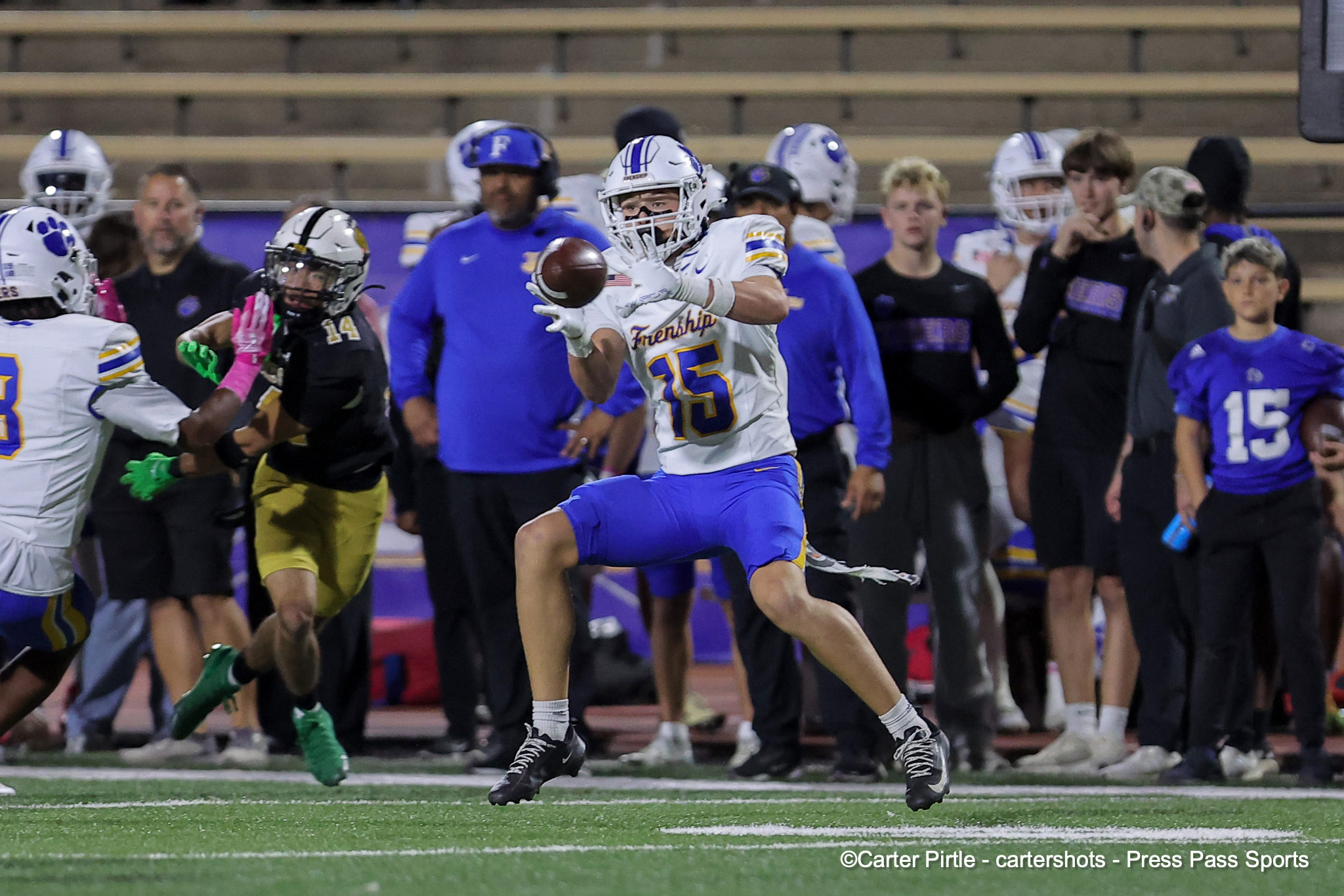 Football player in white and blue (#15) stretches to catch a pass on the sideline with defenders and teammates nearby.