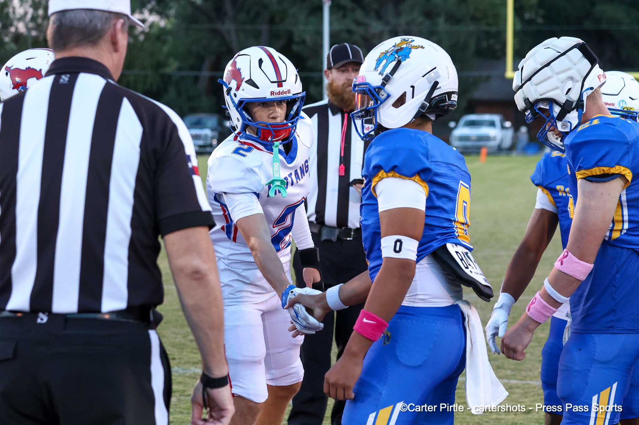 Referee in black-and-white stripes talks to players in blue and white uniforms on a grassy field before a game.