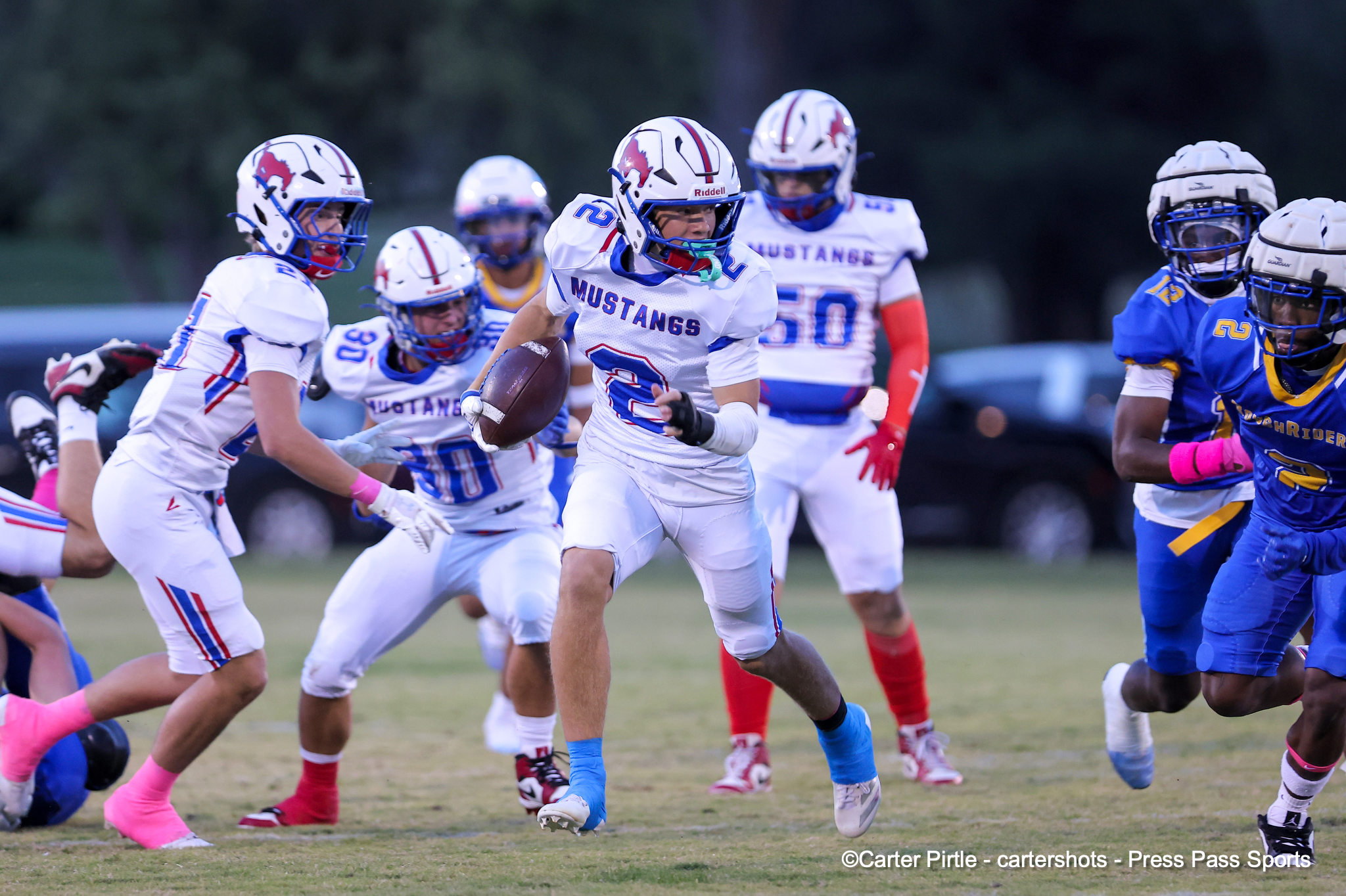 A football player in a white Mustangs uniform runs with the ball as teammates and defenders close in on a grassy field.