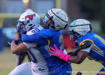 20261023-Claude-BR-FB-004 | Press Pass Sports Rugby players in blue uniforms tackle a white-uniformed opponent during a match, arms and bodies intertwined in a close scrum-like moment.