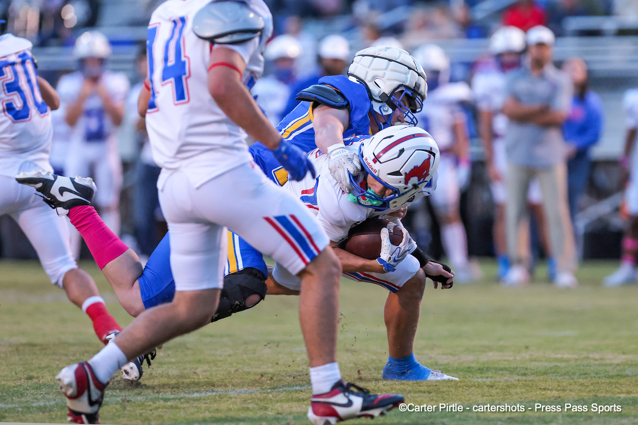 A blue-uniform tackler grips a white-uniform player carrying the ball during a crowded tackle on a football field.