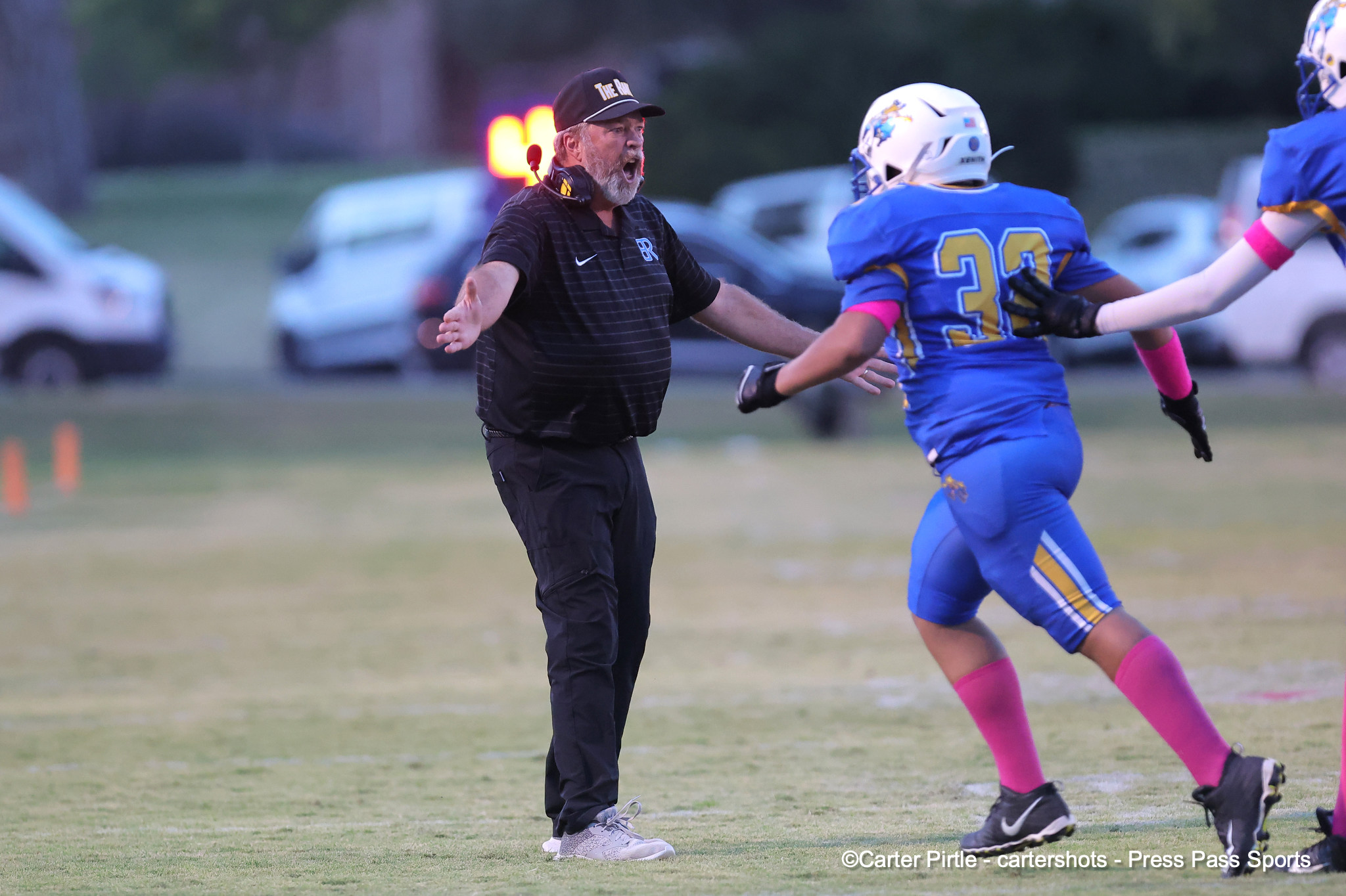 Football coach in black polo and headset shouting at players on a grassy field during a game, blue-uniform teammates running past.