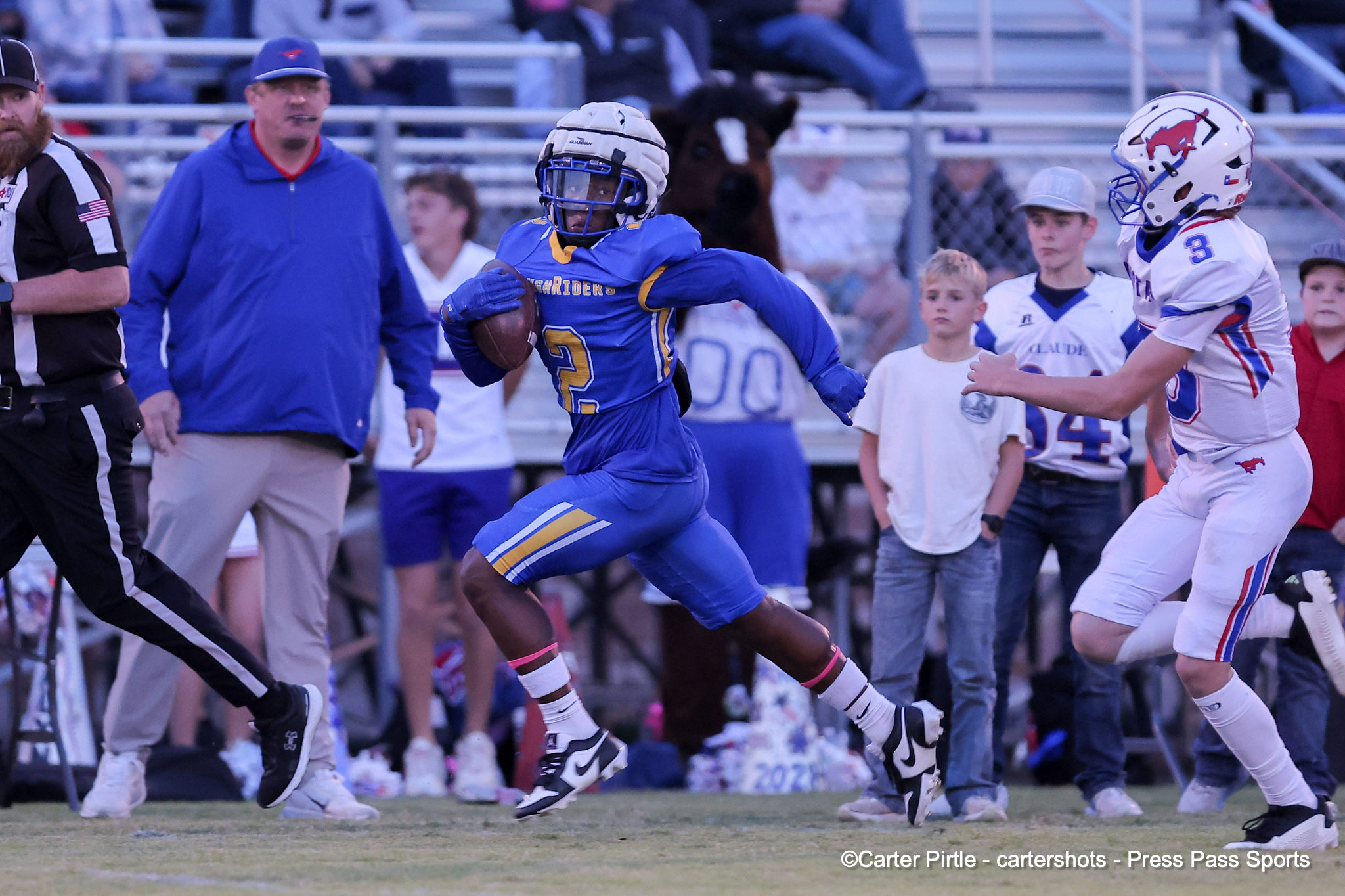 Football player in blue uniform runs with ball while a white-uniform defender pursues; crowd and sidelines in the background.
