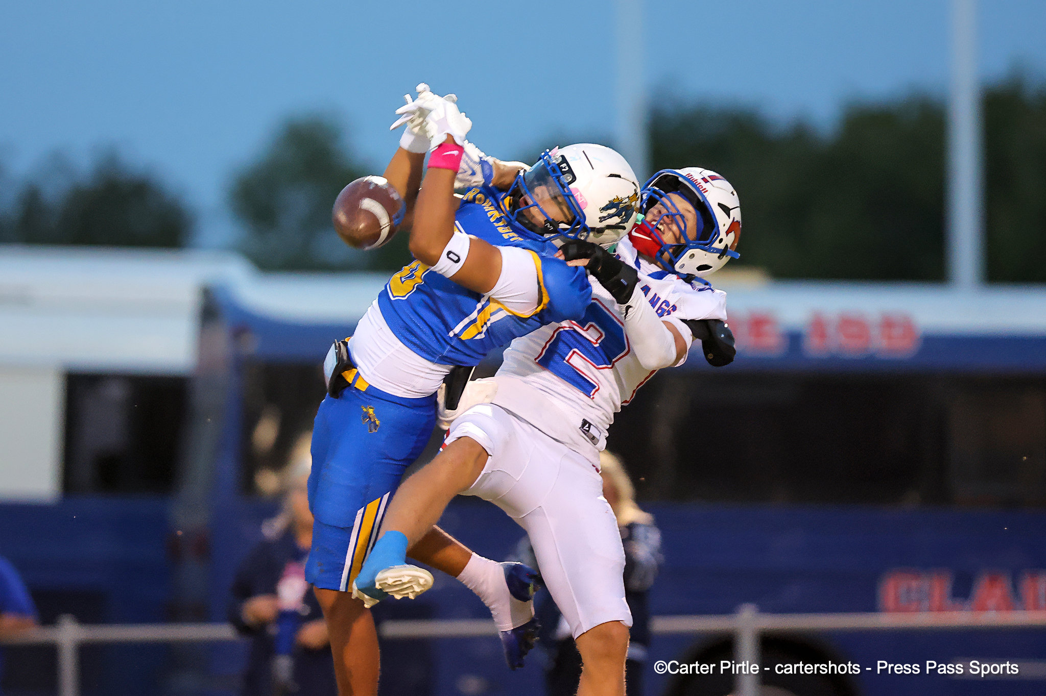 Two football players in blue and white uniforms leap and contest a airborne catch as the ball hovers between them.