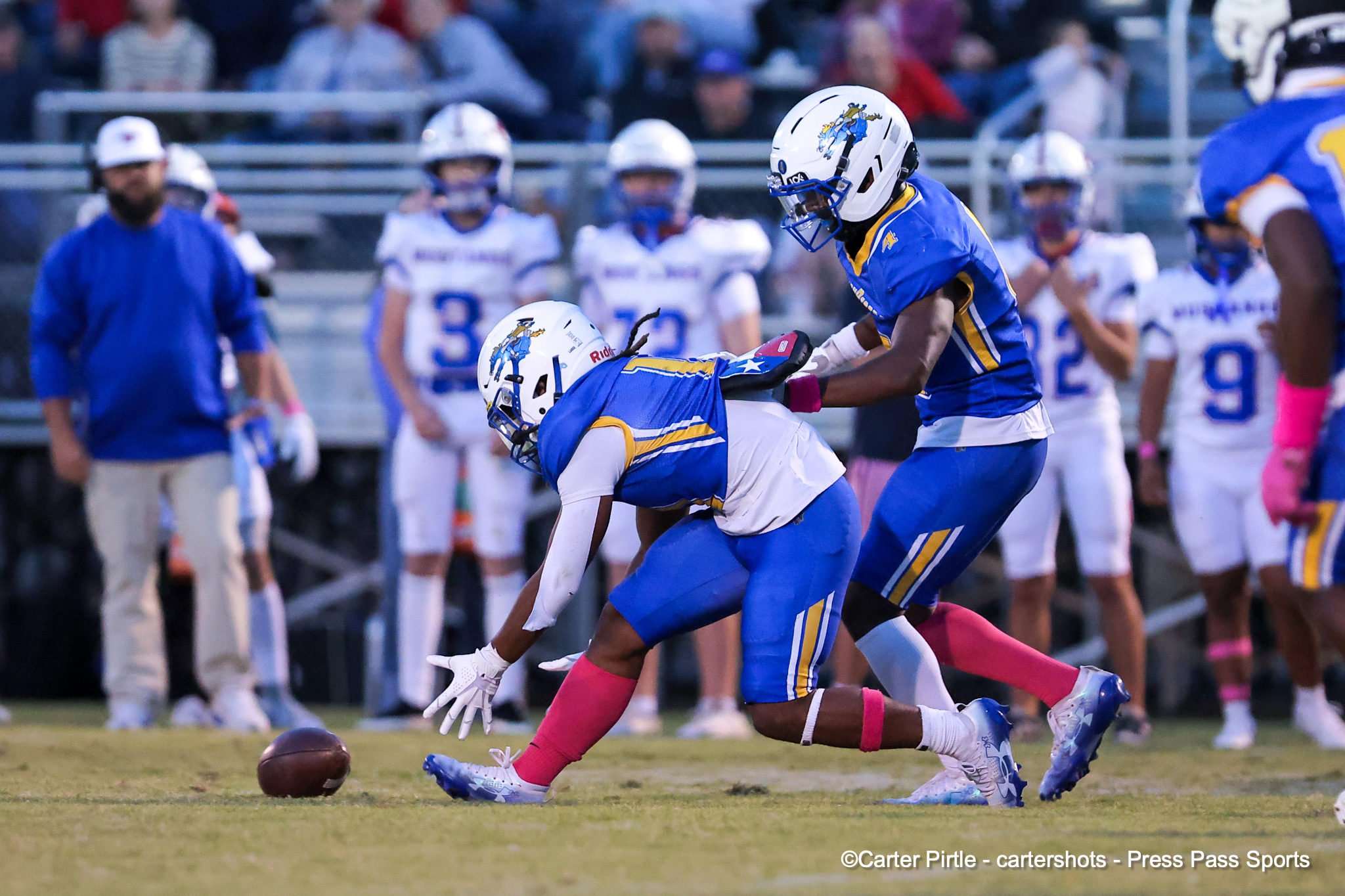 Two football players in blue and yellow uniforms reach for a loose ball on the field as a crowd watches from the stands behind them.