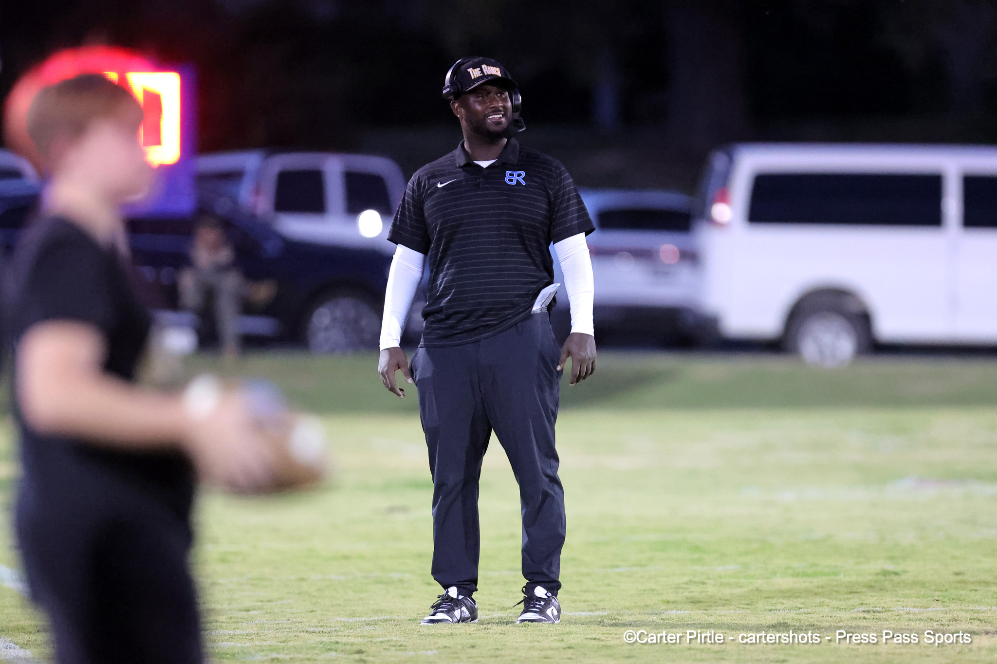 Sports coach standing on a football field in dark striped polo with headset, looking off to the side at dusk or night.