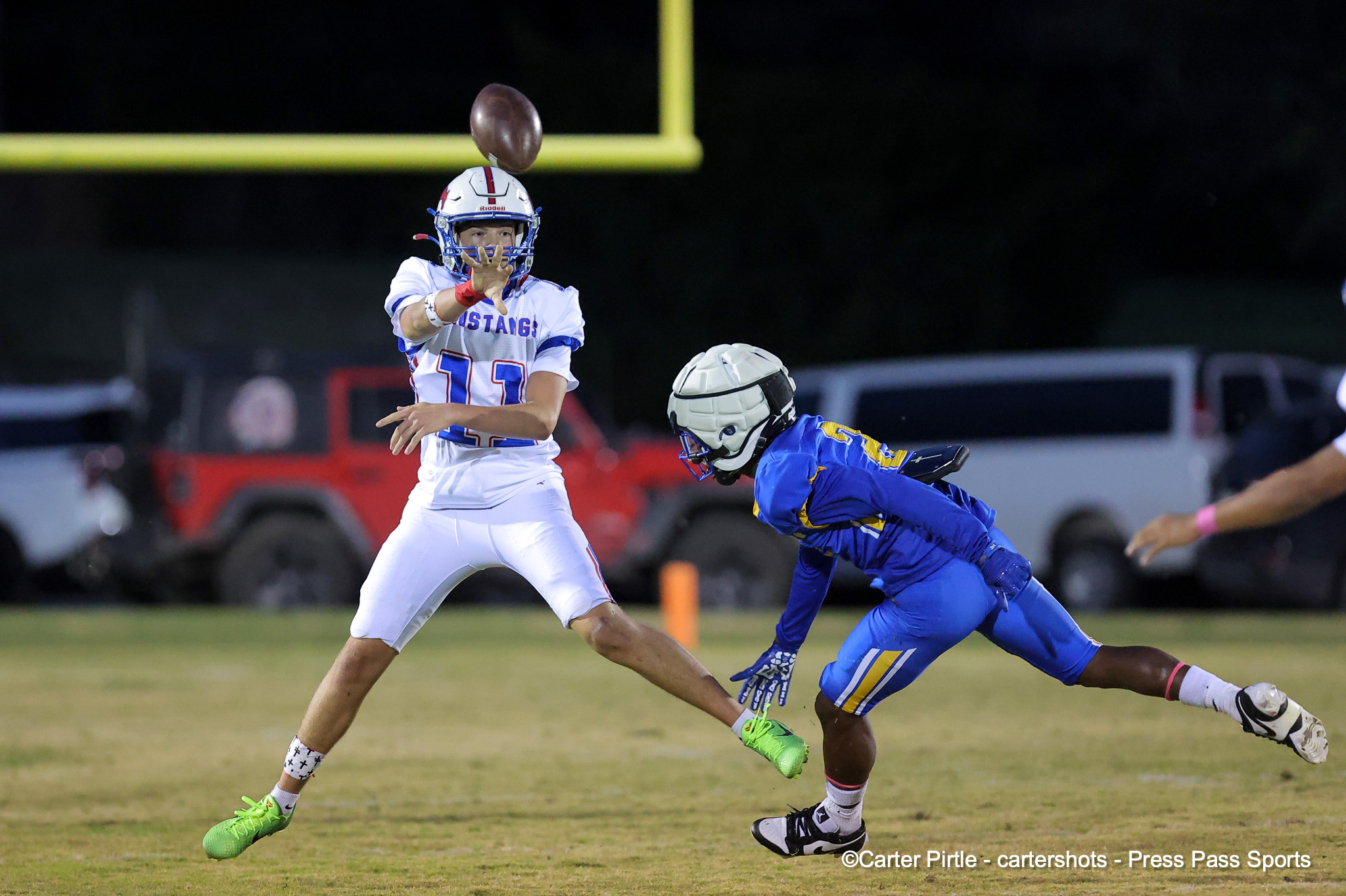 Quarterback in white uniform releasing a pass as a blue defender closes in at night on the field.