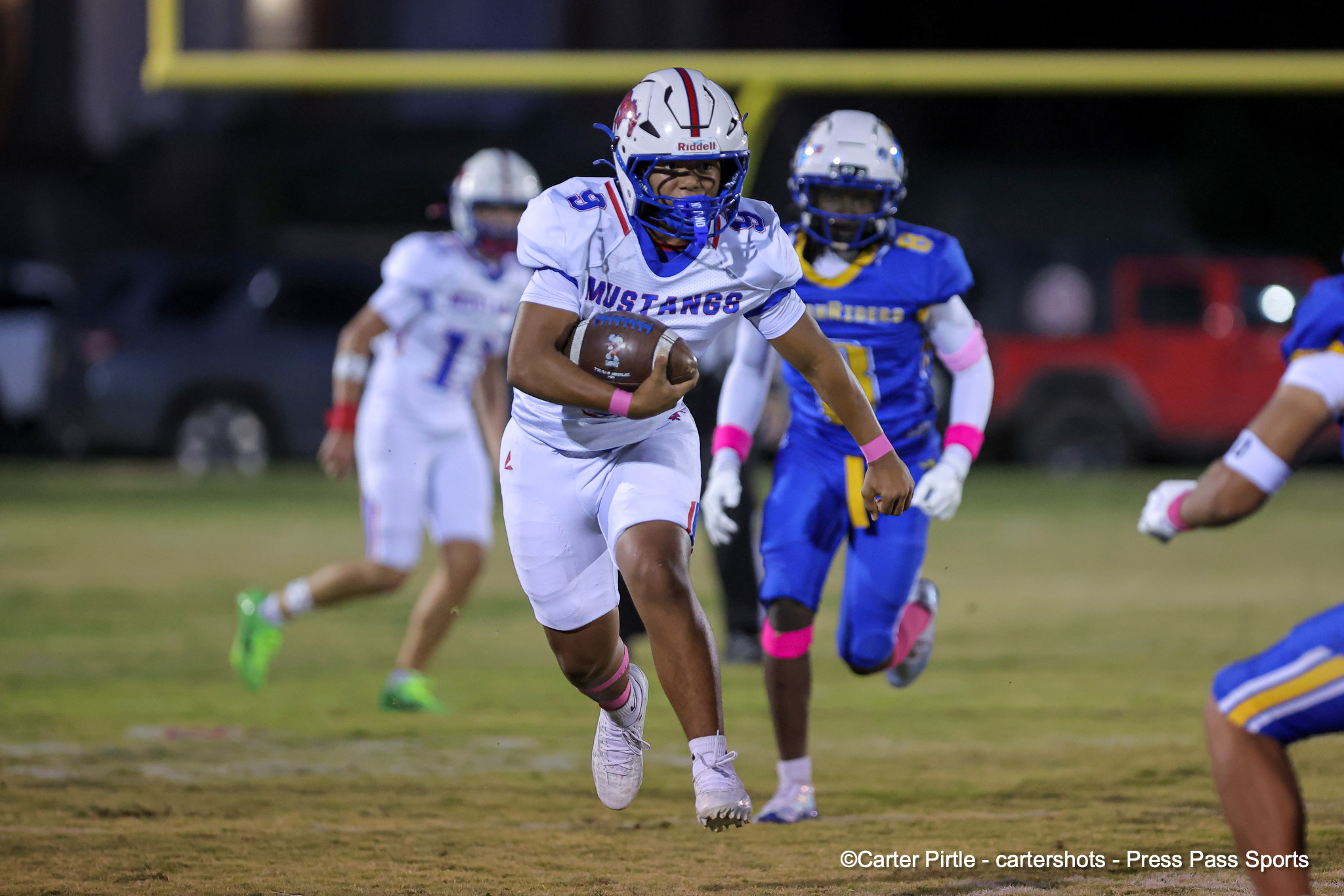 A football player in a white Mustang uniform runs with the ball during a night game, pursued by blue-clad defenders.