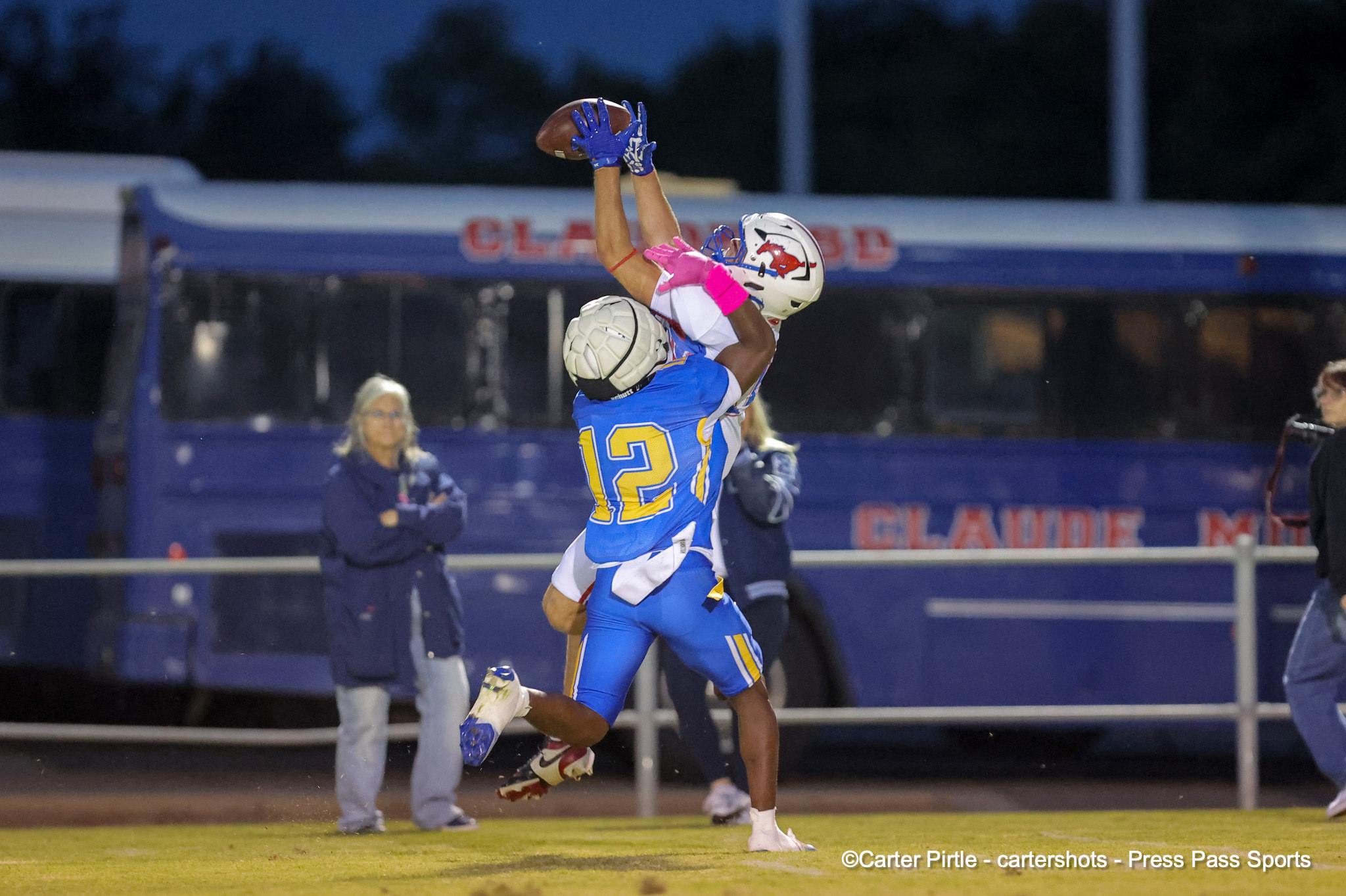 Two football players leap for a pass at night, one in blue with #12 reaching for the ball above the other player in white and pink gloves.