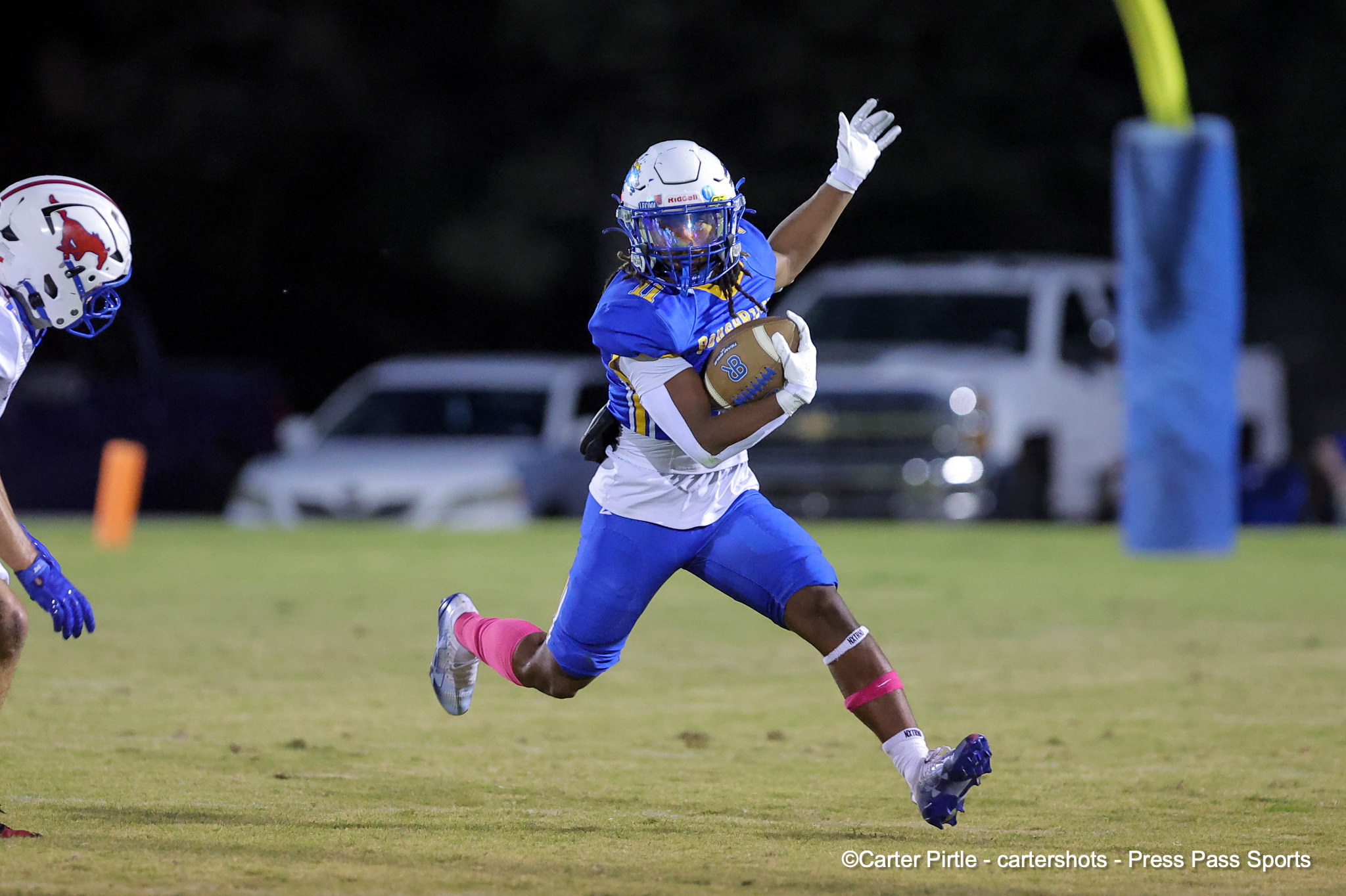 Football player in a blue uniform runs with the ball at night, white gloves, pink socks, defender blurred in the background.