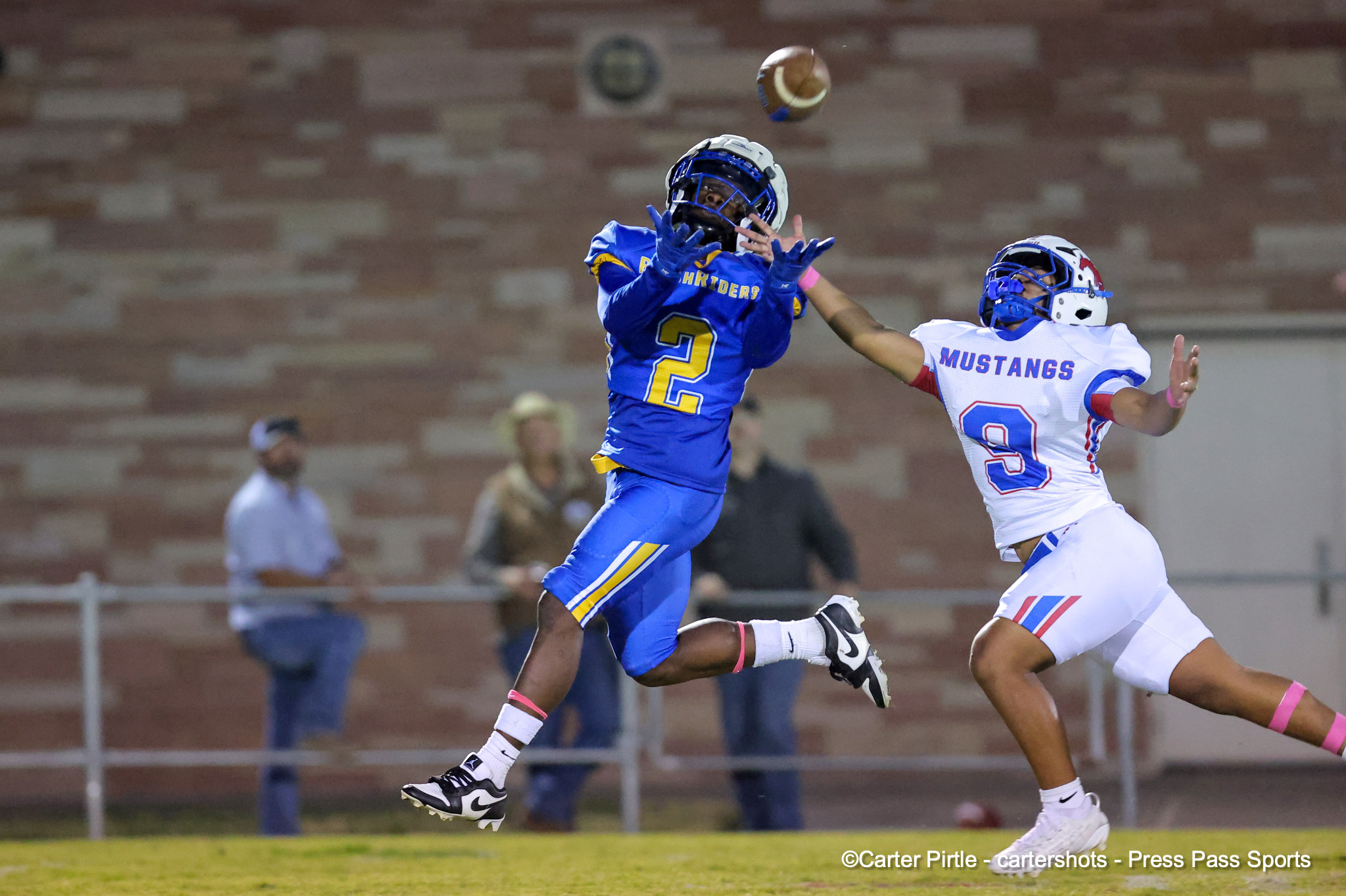 Two lacrosse players leap for the ball in midair, one in blue number 2 and the other in a white'Mustangs' uniform with 0/3 visible.