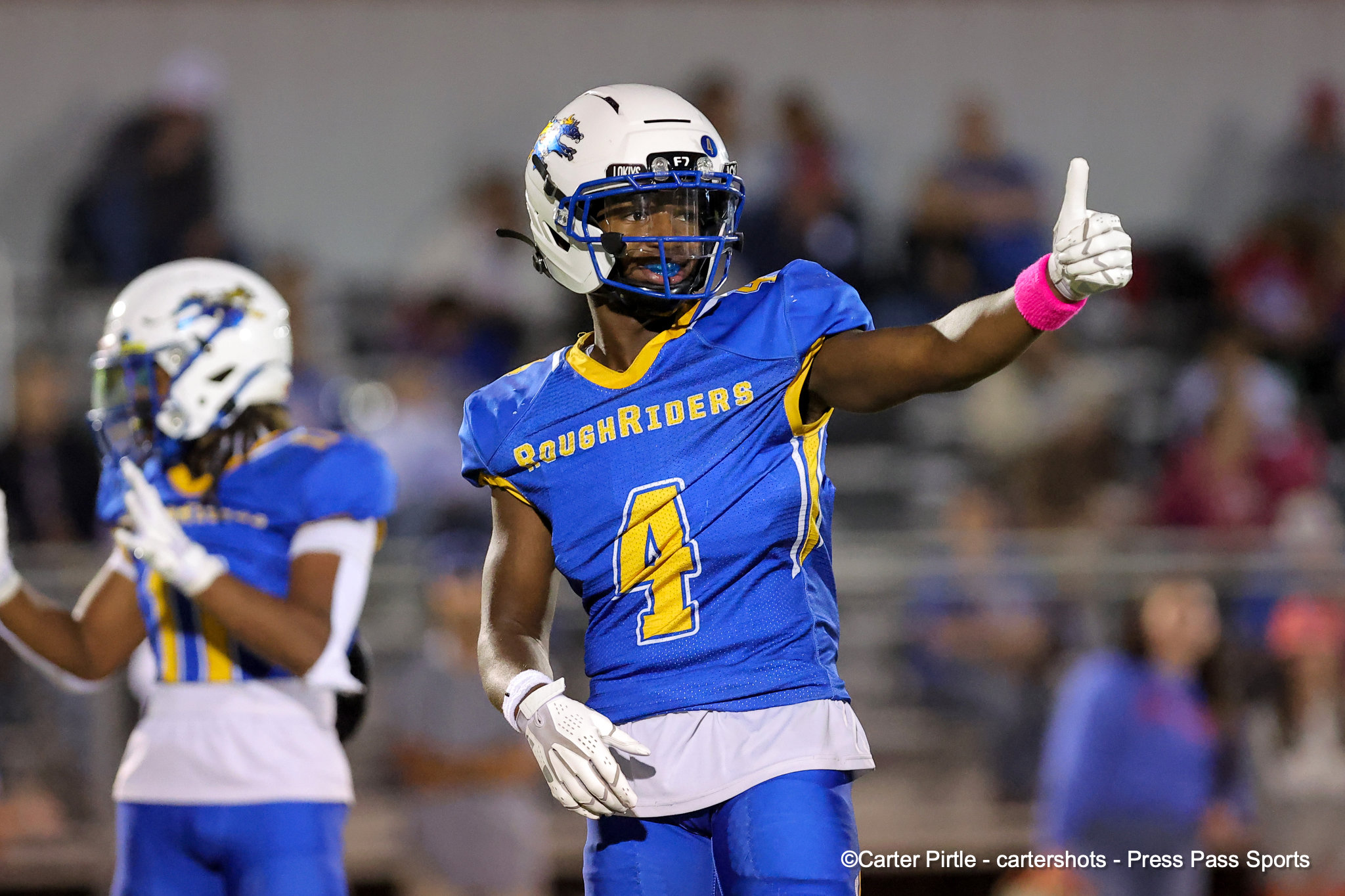 RoughRiders football player in blue and yellow uniform with white helmet giving a thumbs-up on the field, blurred crowd in the background.